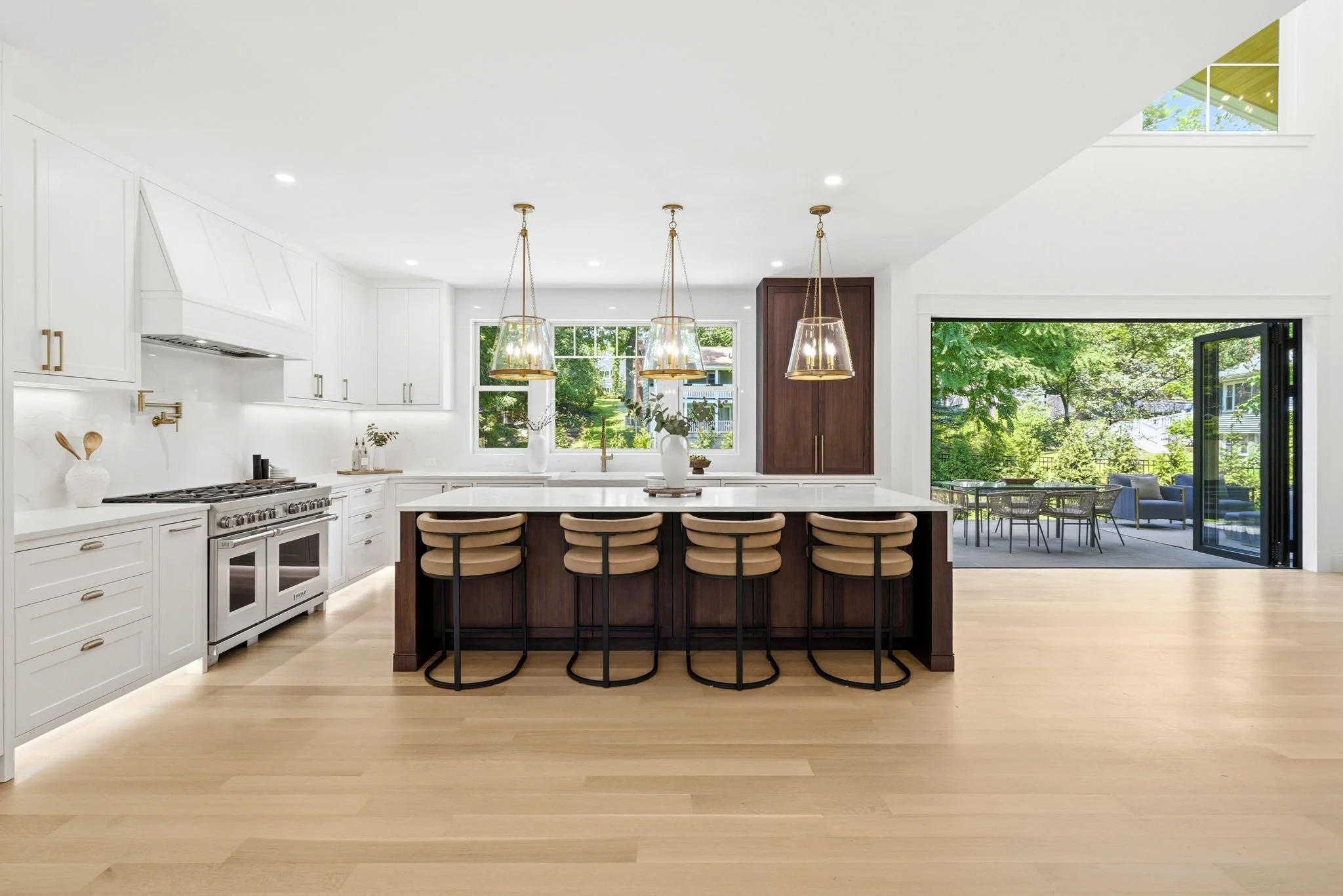 Modern kitchen with white cabinets, a large island with four bar stools, pendant lighting, large windows, and a sliding glass door leading to an outdoor patio with trees and outdoor seating.