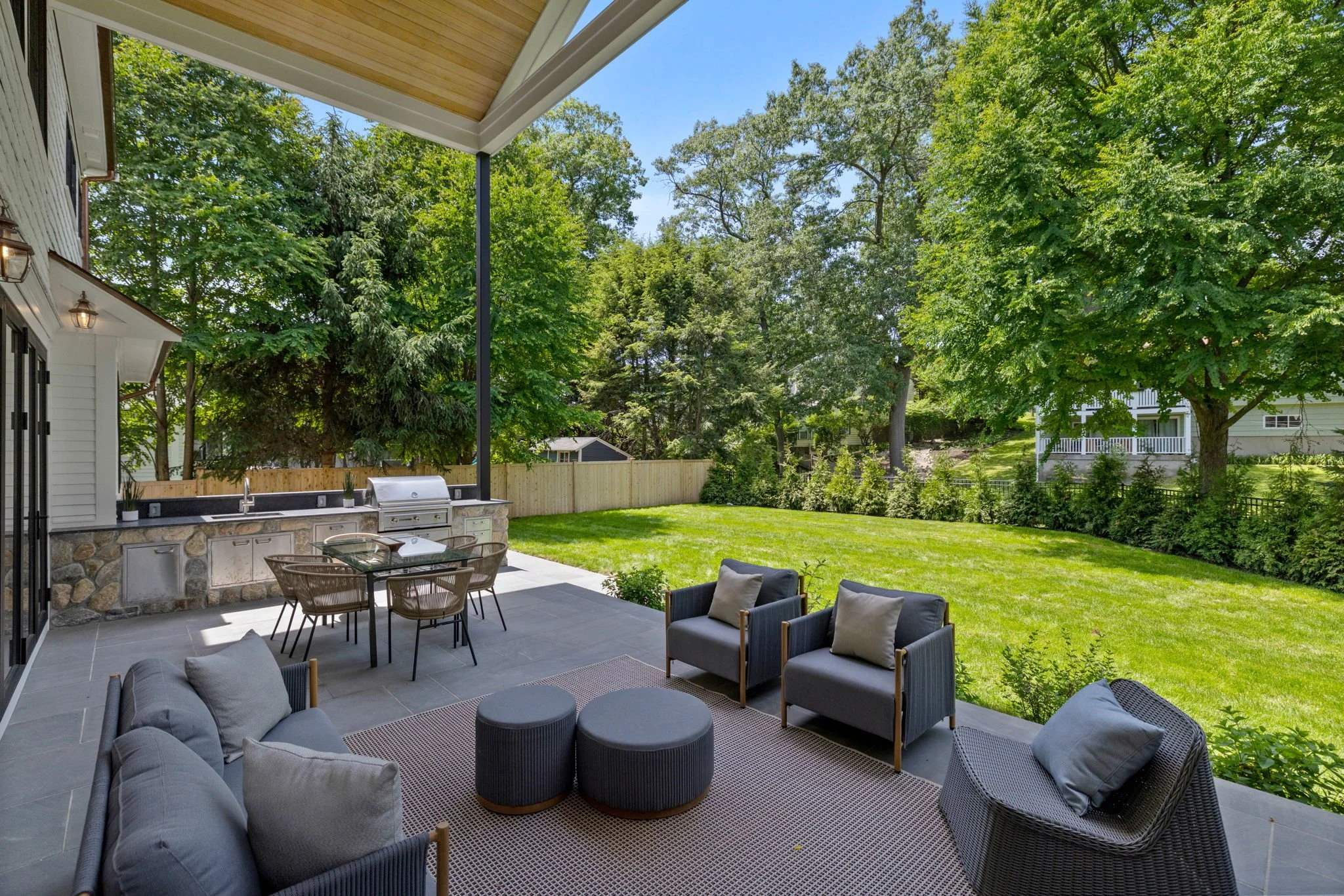 A backyard patio with outdoor furniture including a sectional sofa with cushions, a set of round ottomans, and two armchairs with cushions. There's a built-in outdoor kitchen with a stone base and stainless steel appliances. The patio is paved with l