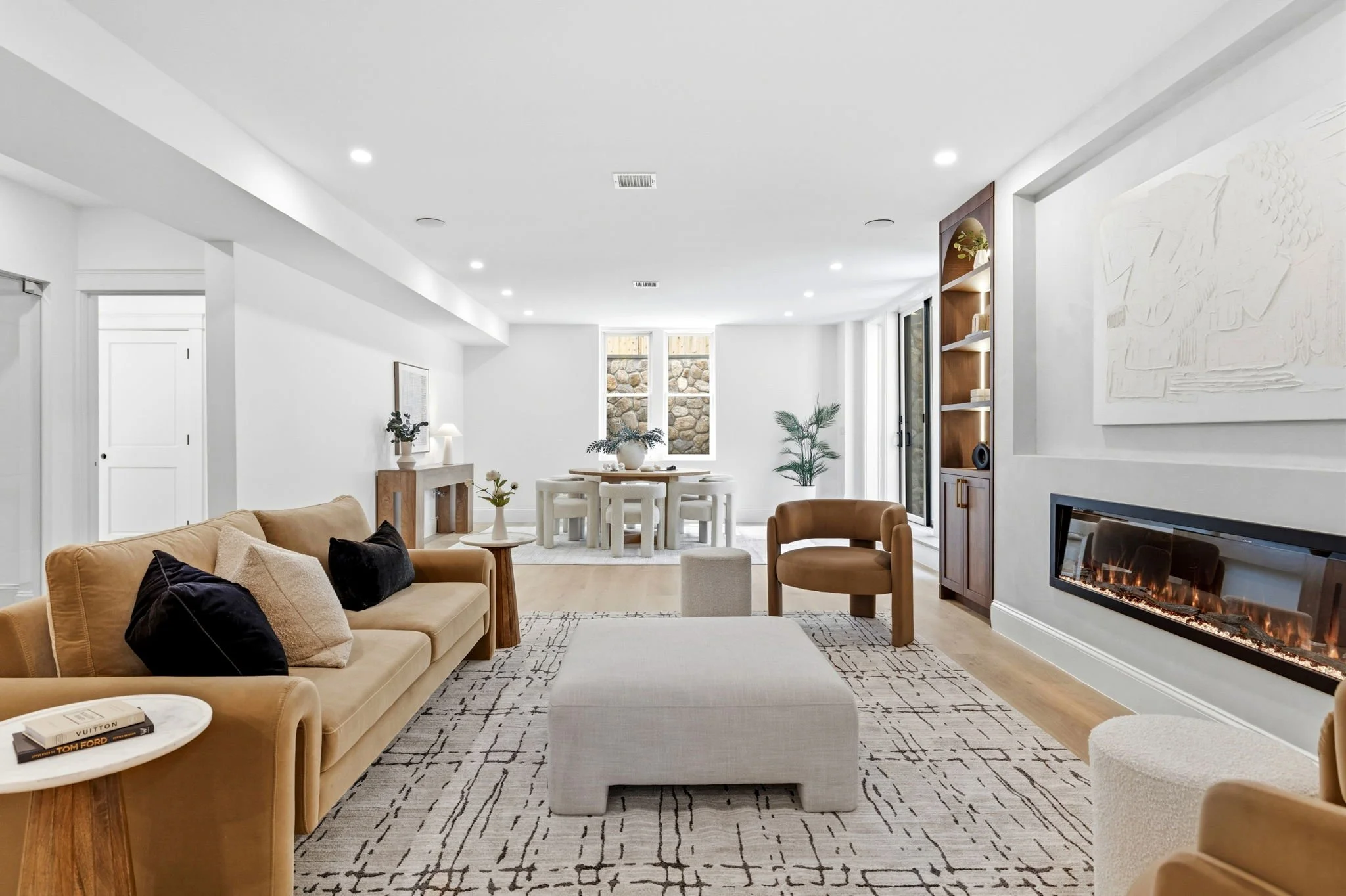 A spacious, modern living room with white walls and ceiling, hardwood floors, beige sofas with black and beige pillows, a light gray ottoman, a patterned rug, a fireplace with a fire, a round side table with books, a wooden console with a table lamp 