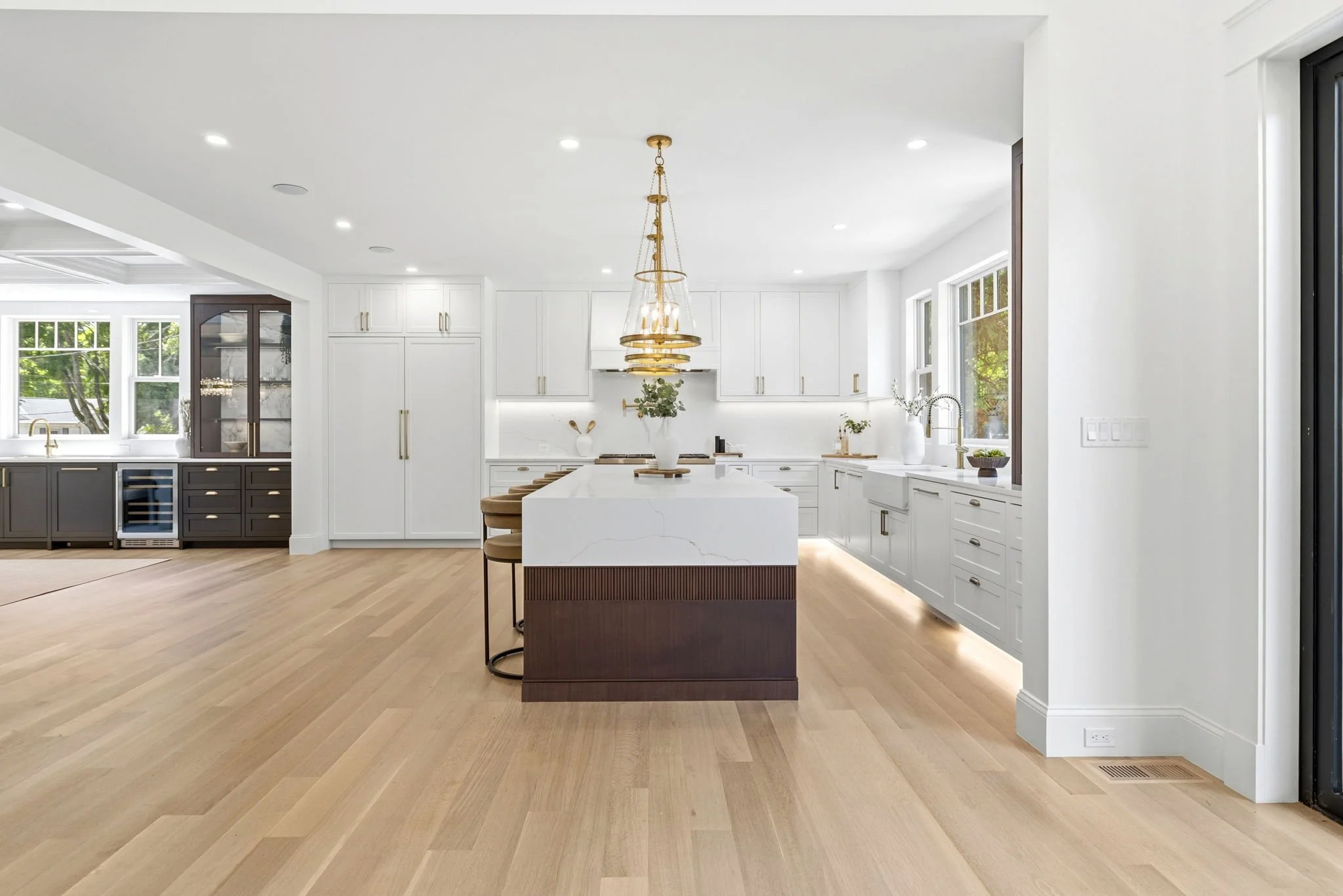 Modern kitchen with white cabinetry and marble island, gold light fixture, large windows, light wood flooring, and black accents.