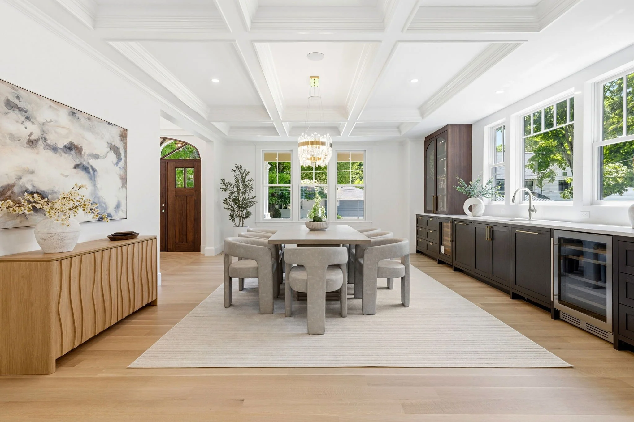 Interior view of a modern, bright dining room and kitchen with large windows, white walls, a white coffered ceiling, a wooden front door, a chandelier, a dining table with upholstered chairs, a sideboard with a vase and decorative object, a built-in 