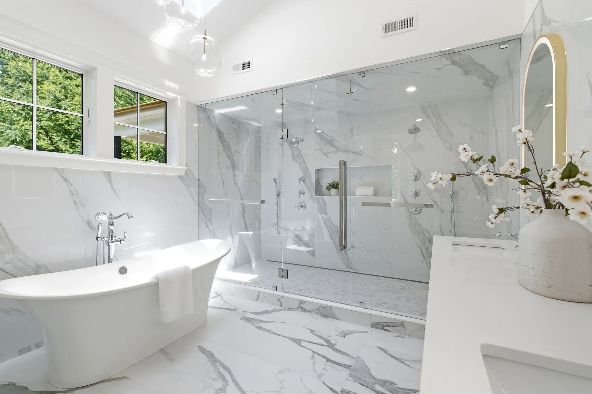 A modern bathroom with white marble walls and floor, a clawfoot bathtub, a glass-enclosed shower, large windows showing green trees outside, a vase with white flowers, and minimalistic decor.