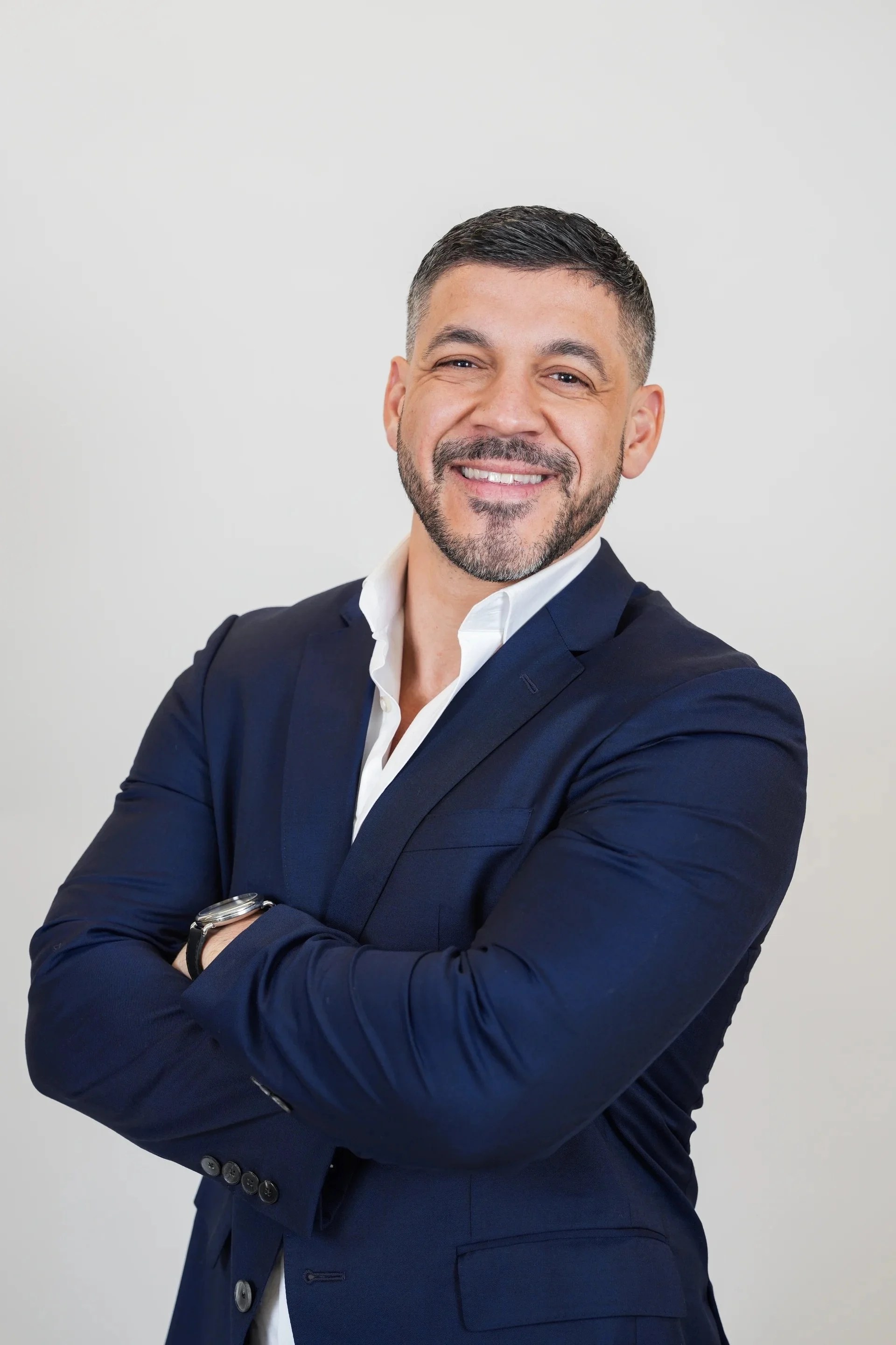 A man with short dark hair and a beard, wearing a navy blue suit and a white shirt, smiling with arms crossed, standing against a plain light gray background.