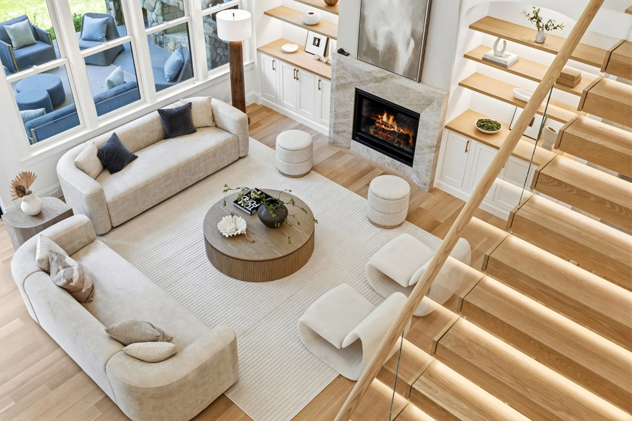 Living room with beige sofas, a round wooden coffee table, fireplace, built-in shelves, large windows, hardwood floors, and a staircase with wooden steps.
