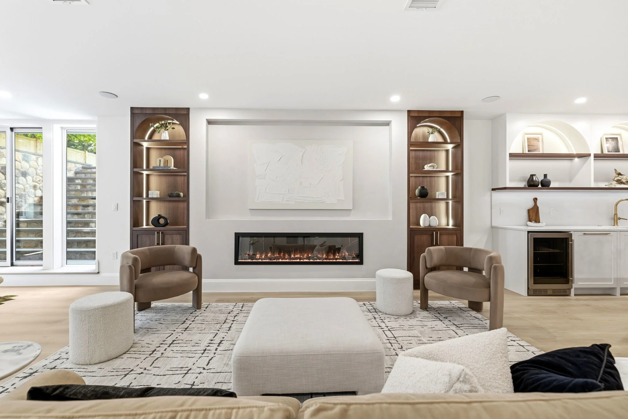 Modern living room with a fireplace, wooden shelves, beige armchairs, a white ottoman, and decorative items on the shelves.