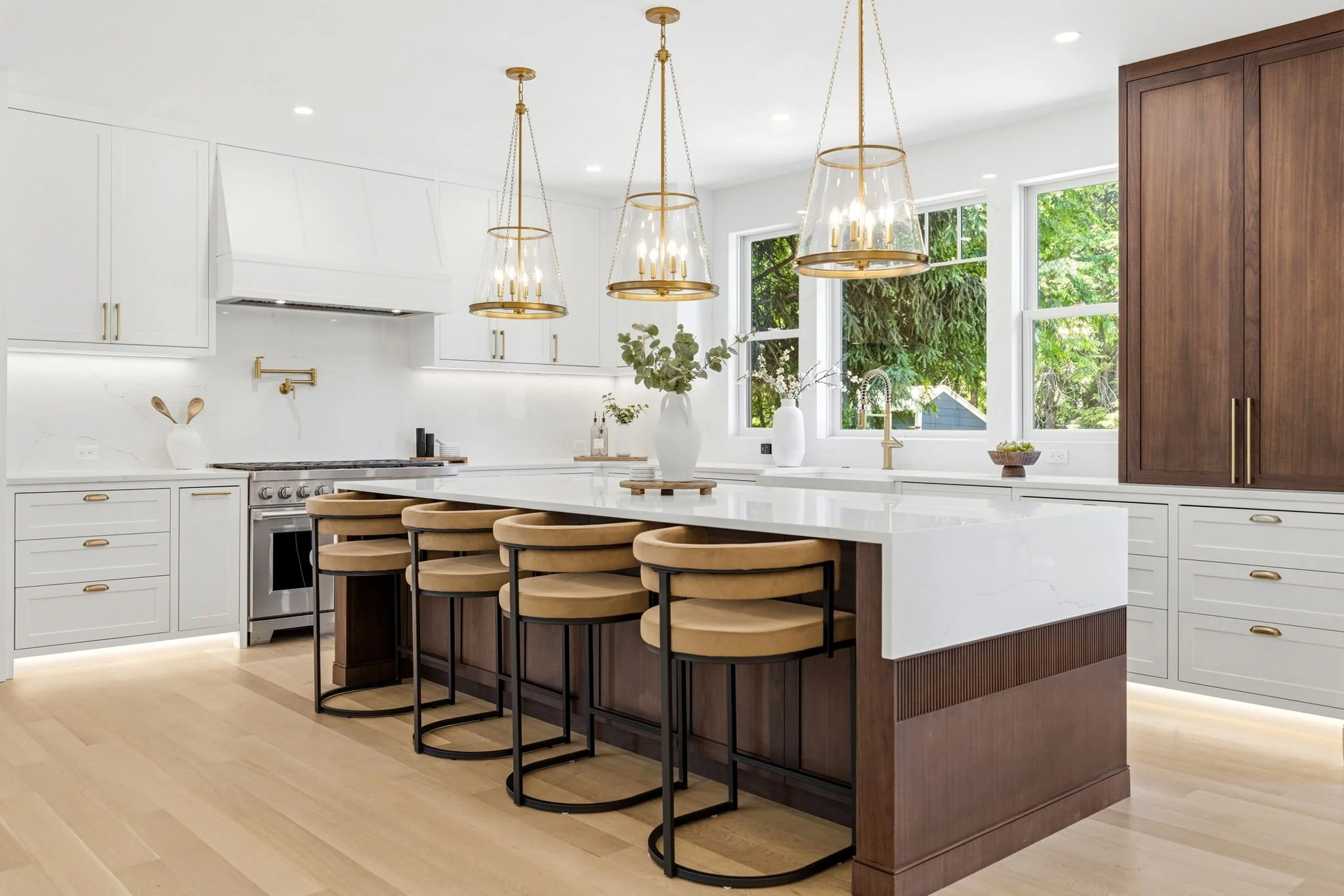 Modern kitchen with white cabinets, a large white island with wooden bar stools, and pendant lights hanging above. Large windows show green trees outside.