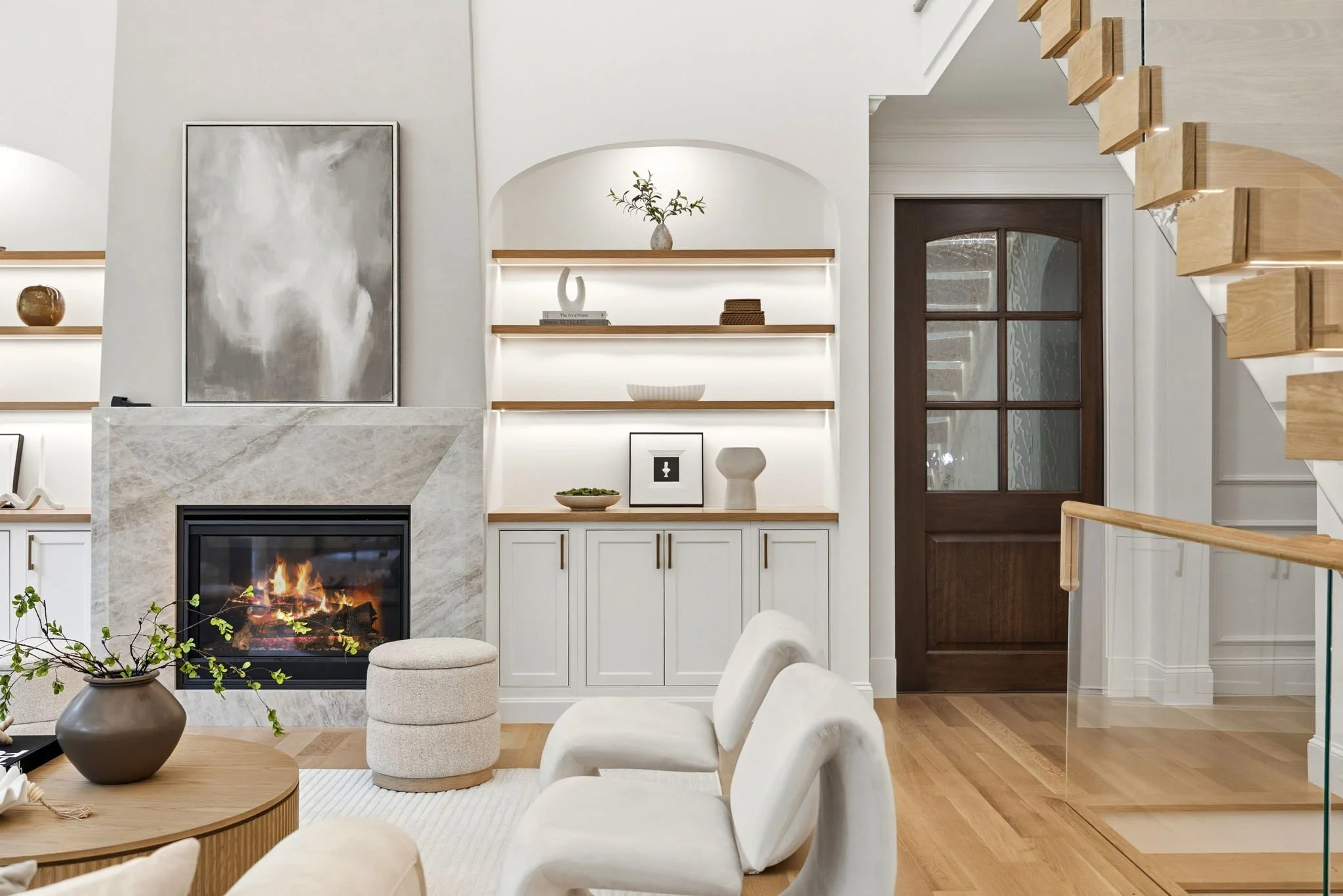 Living room with a modern fireplace, white built-in shelves decorated with vases and books, a dark wooden door with glass panels, staircase with wooden steps, and white chairs around a round wooden table. Potted plant in the foreground.