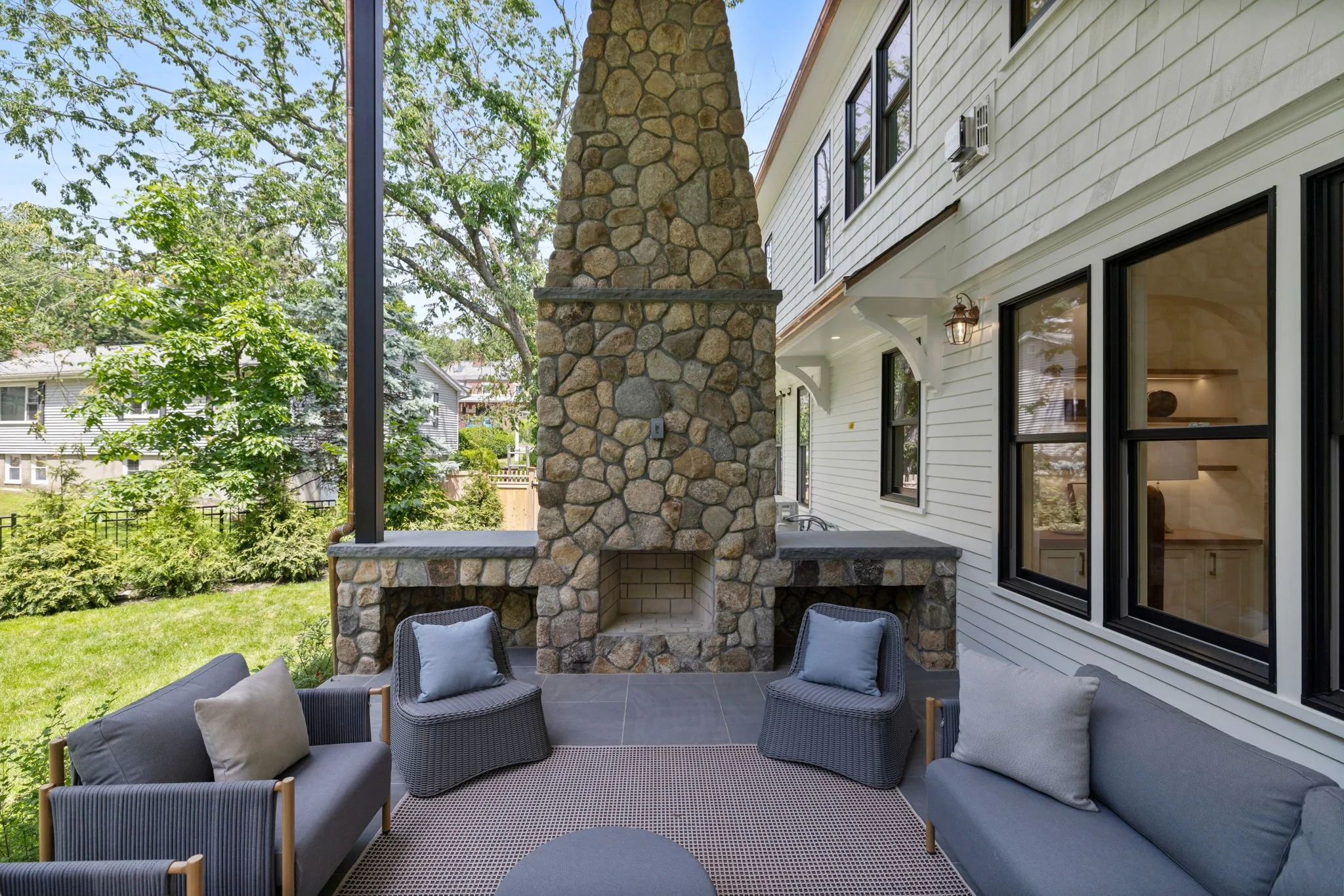 Outdoor patio with gray wicker furniture and beige cushions next to white house with black window frames, stone fireplace, and trees in the background.
