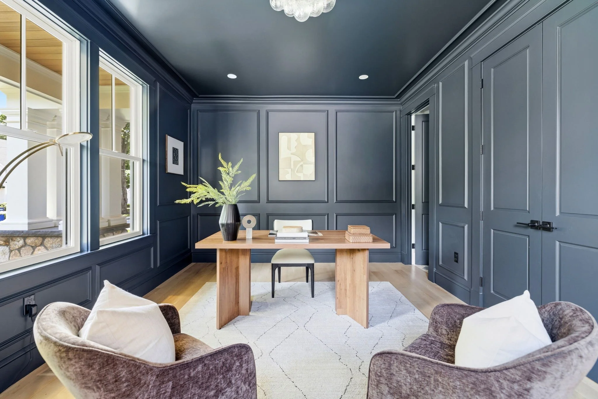 A modern dining room with navy blue paneled walls and large windows. A wooden dining table with a black and cream vase holding green foliage, surrounded by a mix of plush chairs and an armchair. Two pieces of abstract wall art hang on the walls. The 