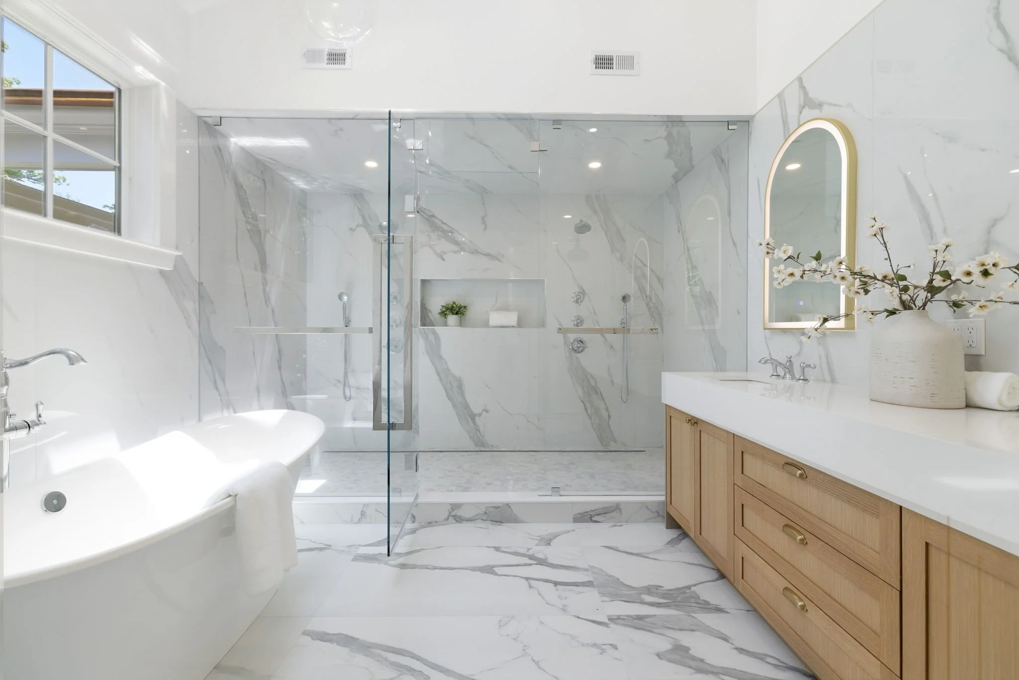 Modern bathroom with white marble walls and flooring, a glass shower with built-in shelves, a white bathtub, a large window, a wooden vanity with a white countertop, a mirror with lighting, a vase with white flowers, and recessed ceiling lights.
