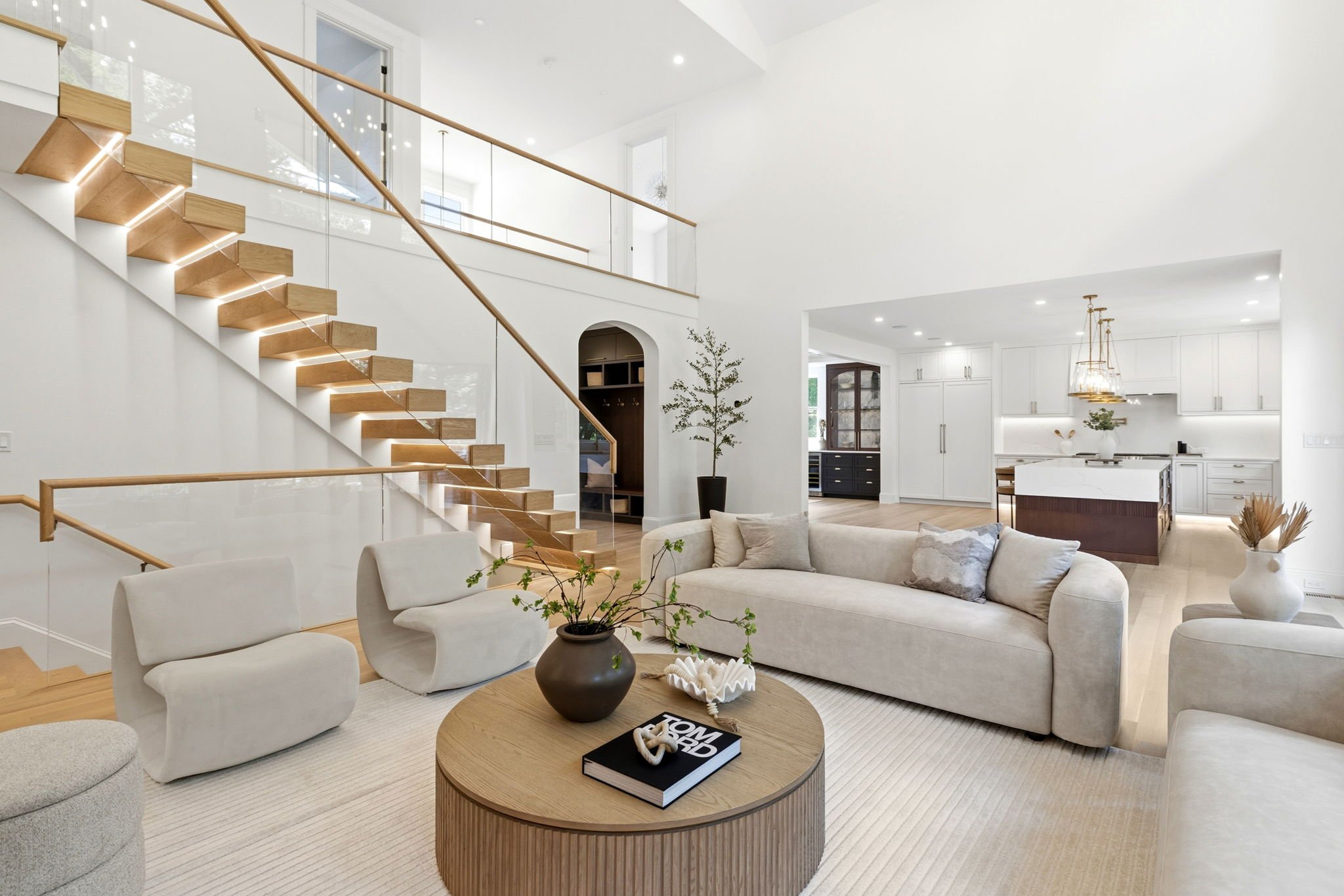 Open-concept living room with white walls and ceiling, featuring a staircase with wooden steps and glass railing, a white sectional sofa, a round wooden coffee table, and modern decor including a potted plant and books.