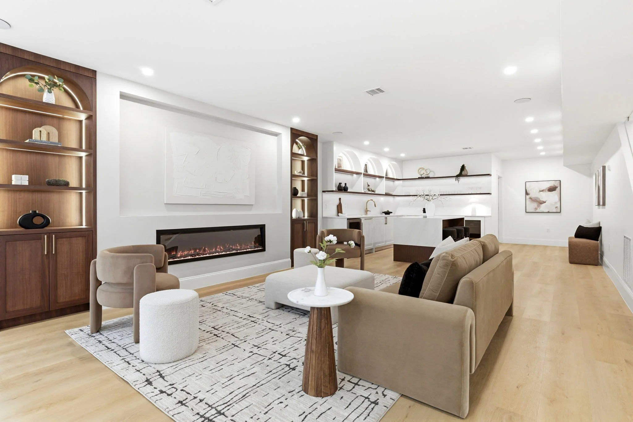 Modern living room with beige sofas, a white round coffee table with flowers, a fireplace, and built-in shelves with decorative items. Light wood flooring and white walls with minimalistic artwork.