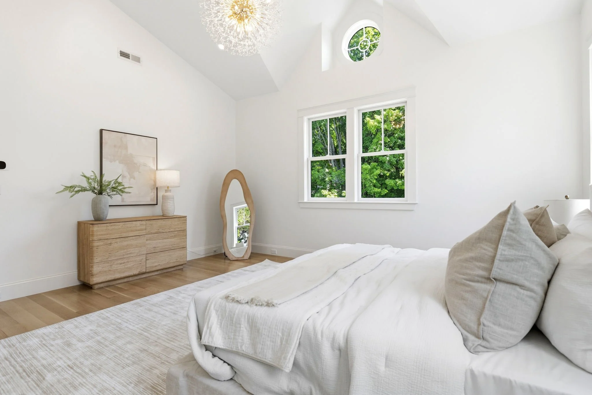 A bright, minimalistic bedroom with white walls, large windows showing green trees outside, a wooden dresser with a plant and lamp, a full-length mirror, and a neatly made bed with beige pillows and white linens.