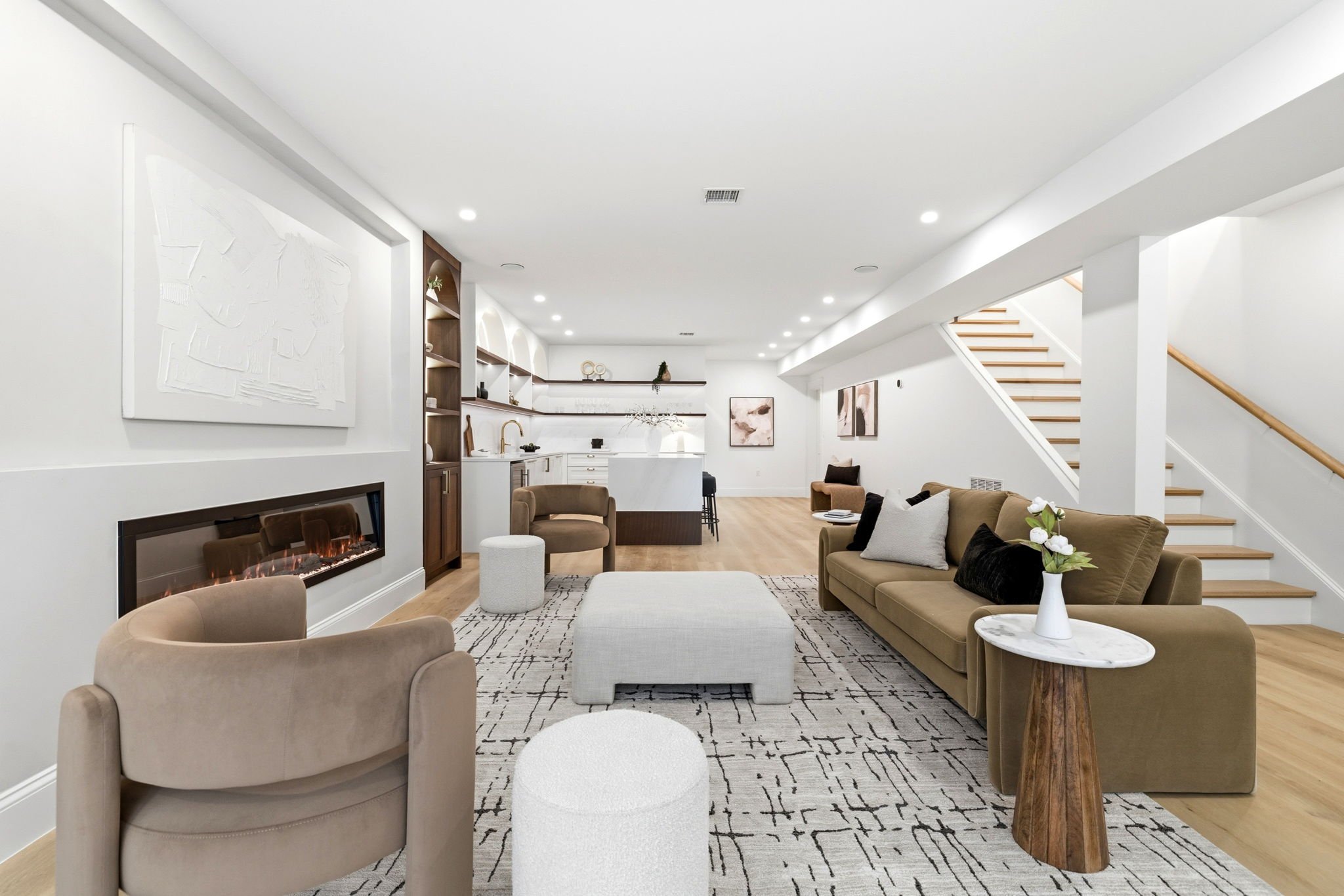Interior of a modern, open-concept living room with white walls, hardwood floors, a beige sofa, armchair, black and white patterned rug, fireplace, artwork, and staircase with wooden handrail.