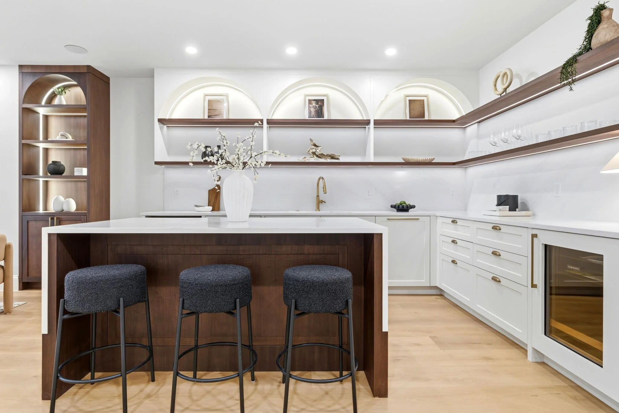Modern kitchen with white cabinets, wooden shelves, a central island with dark stools, and decorative objects including vases, artwork, and glassware against a white wall with wood accents.