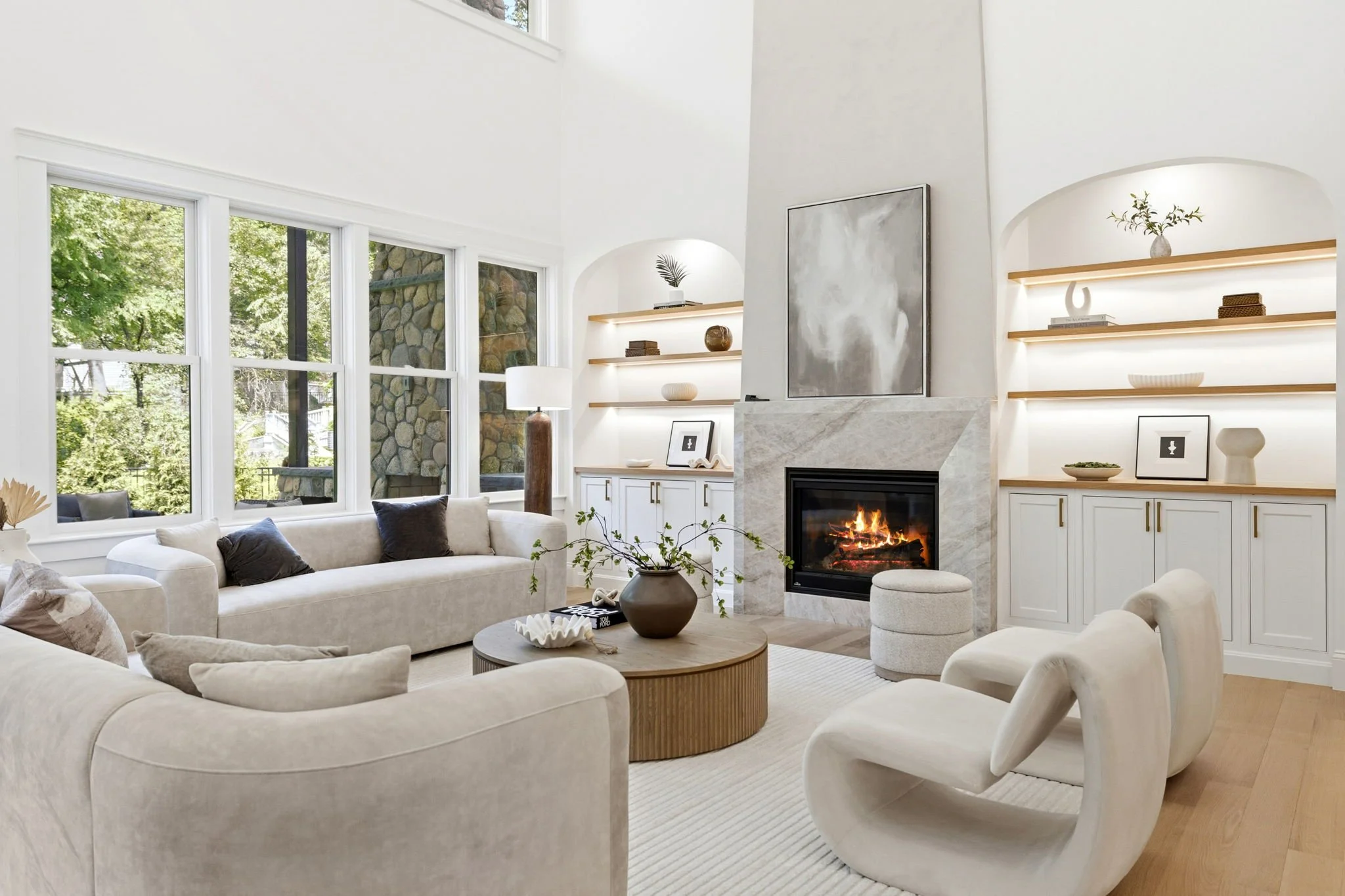 Bright living room with large windows, beige sofas, a round coffee table, a fireplace with a fire, and built-in shelves with decorative items.