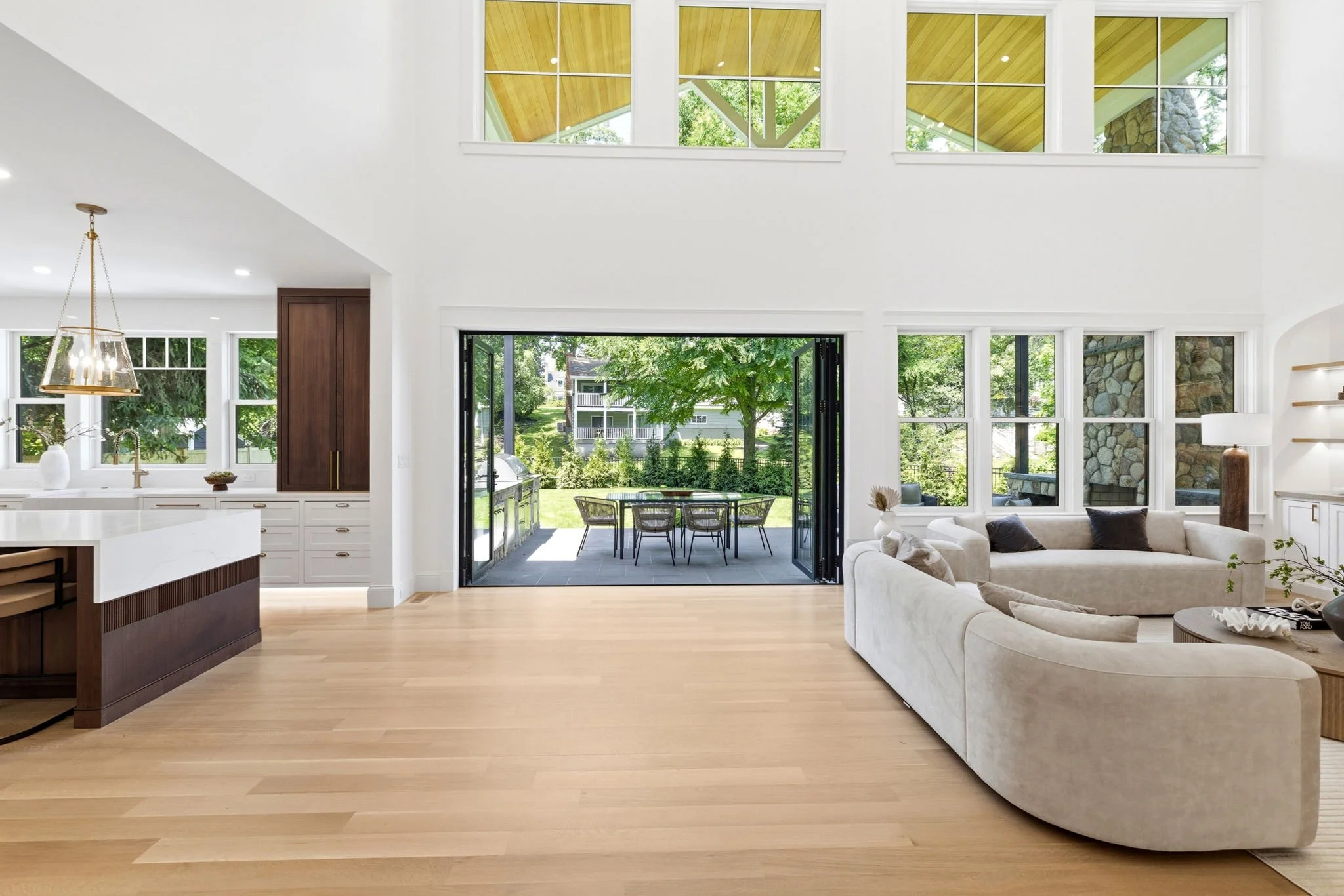 Bright open-concept living space with large windows, a sliding glass door leading to an outdoor patio, and a view of greenery outside. The interior includes a white sofa and a kitchen island with a gold pendant light.