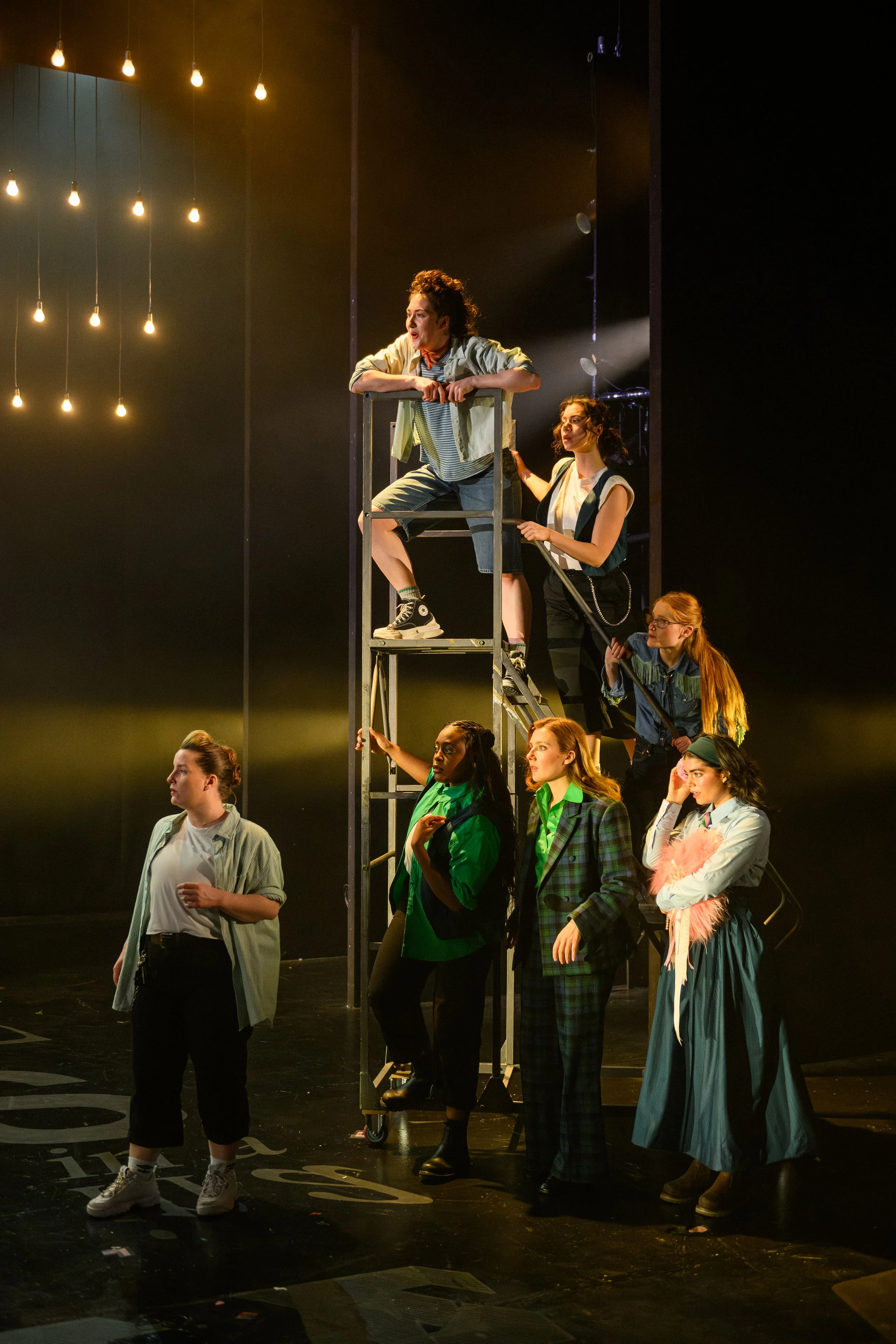 The image depicts a stage performance with a group of actors. A person is standing in the center, talking, while others are positioned around or on a ladder-like structure. The background is dark with hanging light bulbs, creating an atmospheric, cinematic setting.
