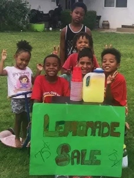 Group of children selling lemonade outside, with a green sign reading 'Lemonade Sale' and bottles of lemonade on the table.