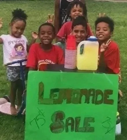Group of children standing behind a green lemonade sale sign outdoors.