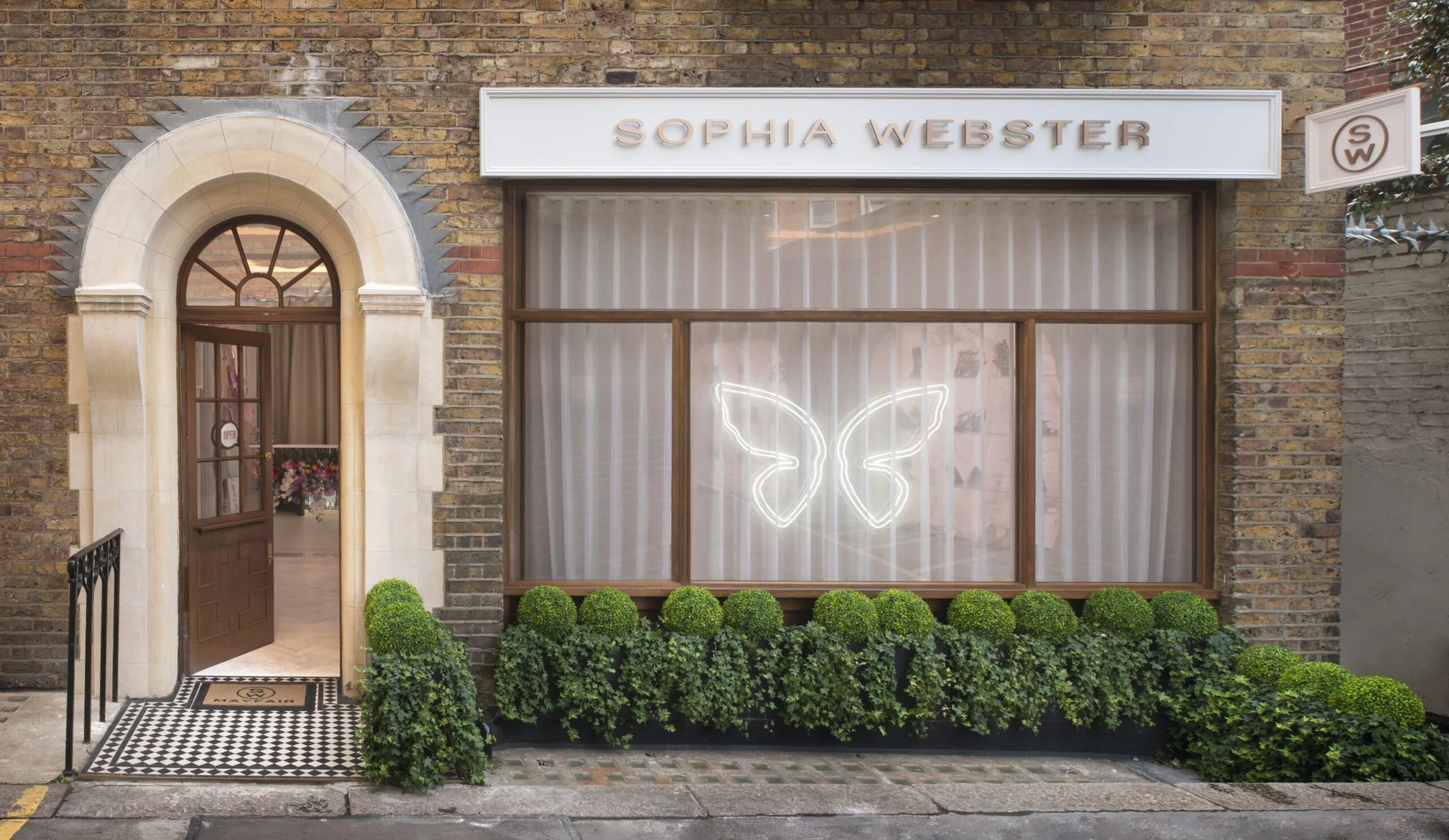 Storefront with sign reading 'SOPHIA WEBSTER', large window with butterfly neon sign, and a flower shop visible through the archway entrance. Front garden with trimmed bushes and checkered floor mat.