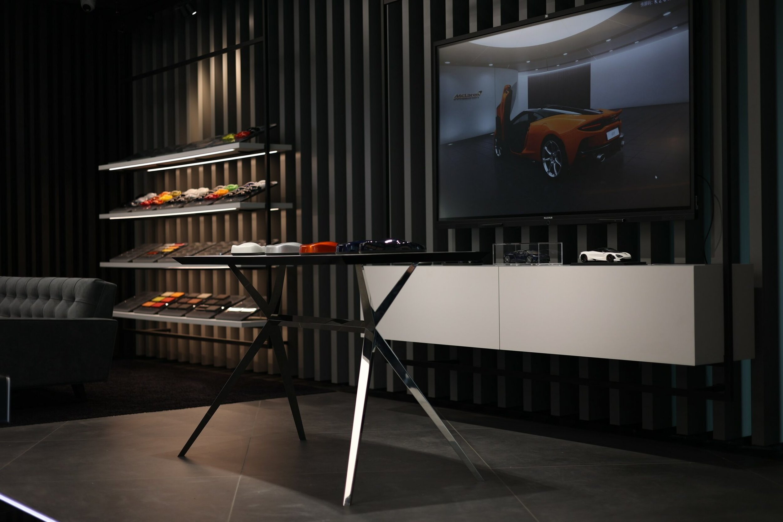 Showroom with scale model cars, color samples, and a large display screen showing a McLaren sports car in an upscale showroom