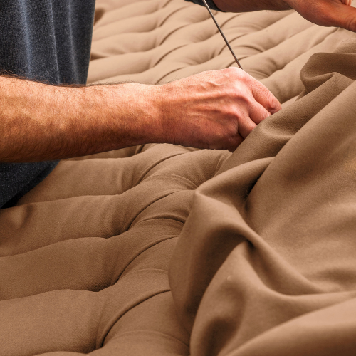 A person is sewing or repairing a beige, tufted fabric surface using a needle and thread.
