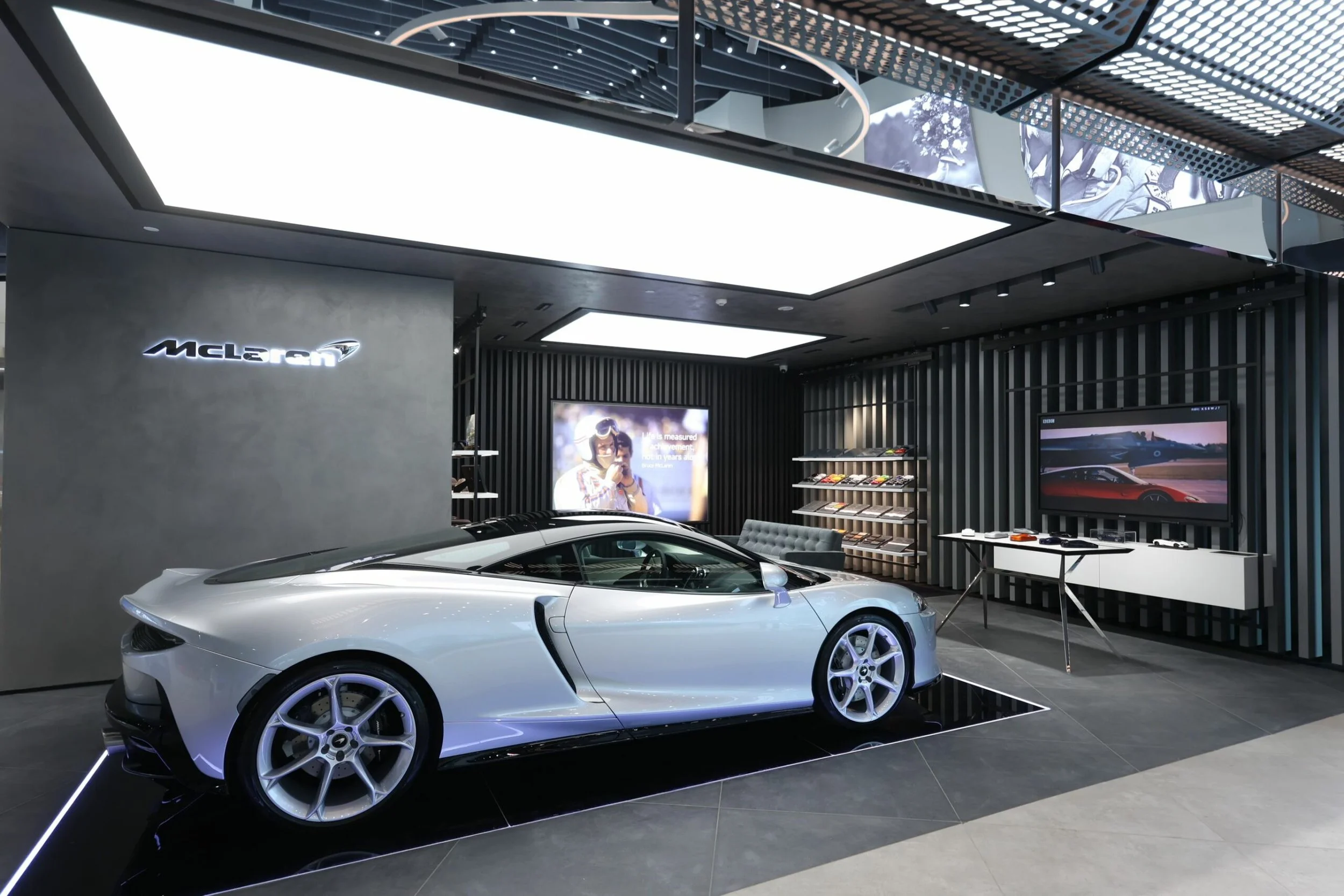 A sleek white luxury sports car on display inside a modern McLaren showroom with digital screens and contemporary decor.