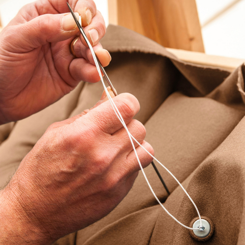 A person threading a needle with white thread through a button on brown fabric.