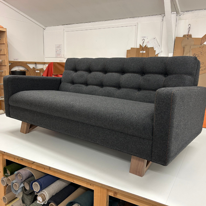 Dark gray upholstered sofa with wooden legs in a workspace or warehouse setting.