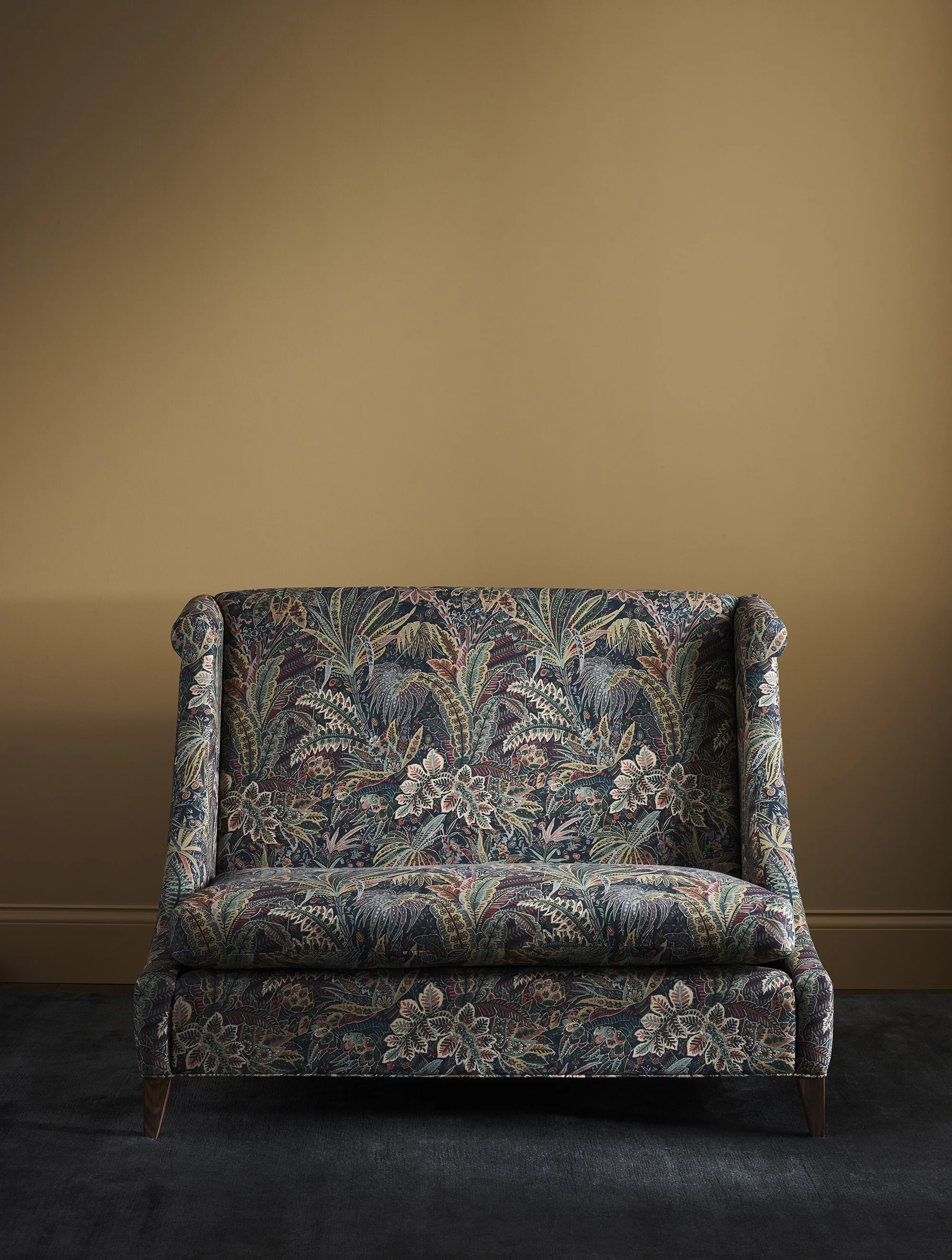 A vintage-style upholstered sofa with a colorful floral pattern, positioned against a plain light brown wall, on a dark carpeted floor.