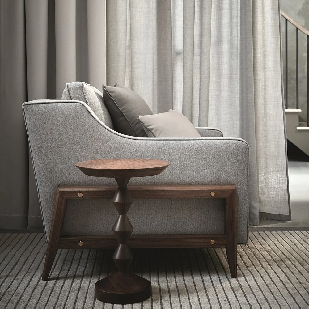 A living room scene with a gray fabric sofa and decorative pillows, a wooden side table, and a backdrop of curtains and a staircase in the background.