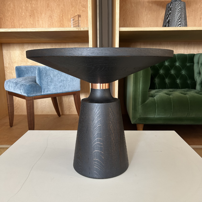 Black round Nicole side table with a conical base and a circular top, placed on a white surface in front of a wooden wall with shelves and two chairs, one green velvet and one blue fabric.