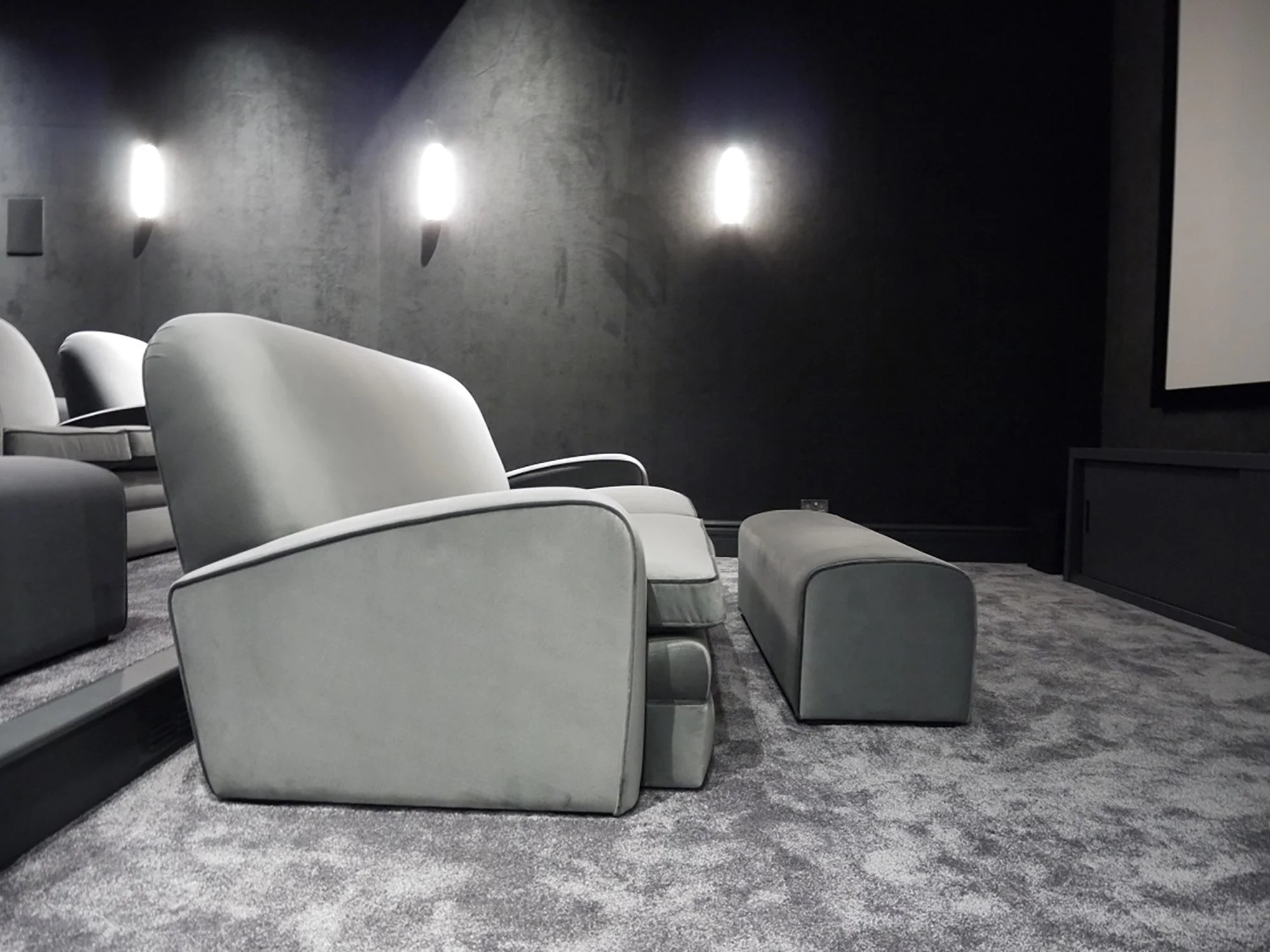Modern home cinema with four white armchairs on a gray carpet, dark wall with three wall-mounted lights, and a large white screen or panel on the right side.