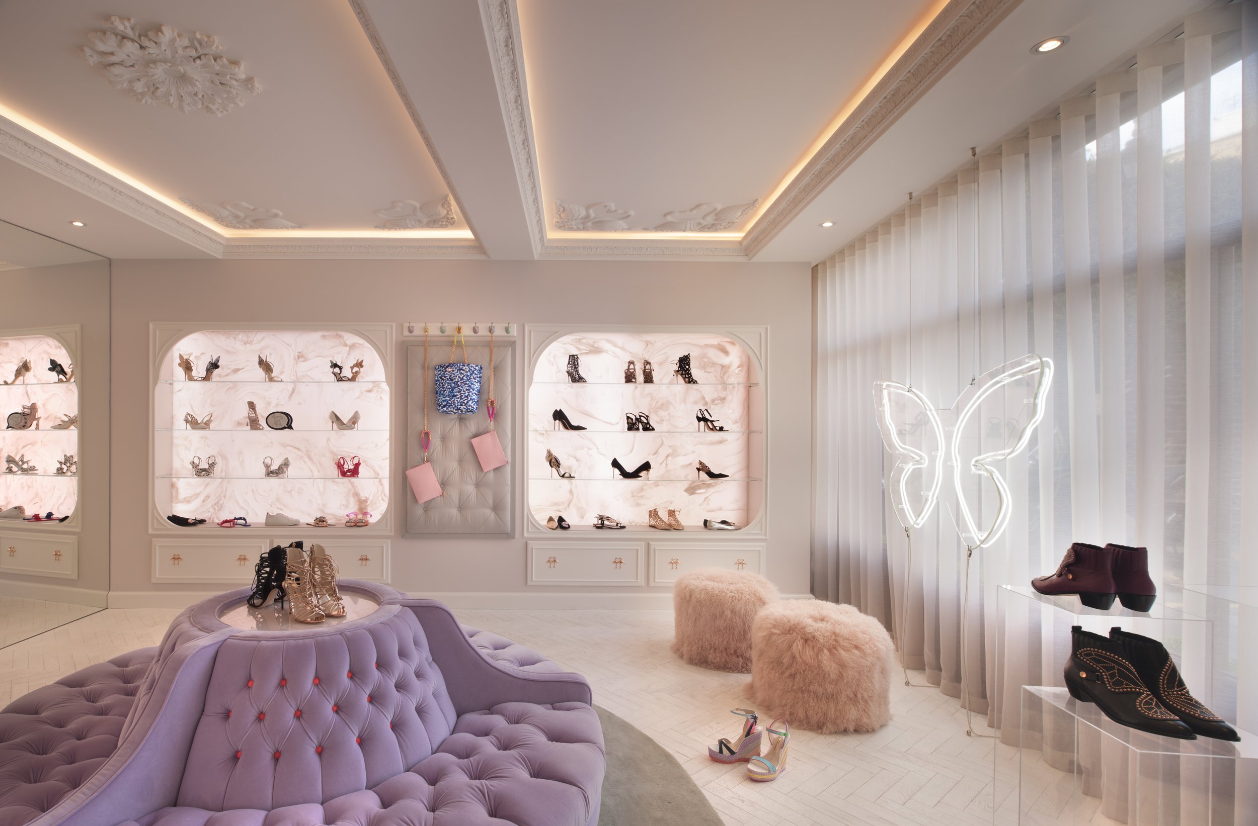 A boutique shoe store with illuminated pink and marble display shelves showcasing various high heels, sandals, and boots. The store has a purple tufted sofa, a plush pink stool, and a butterfly-shaped neon light on the right side, along with floor-length white curtains.