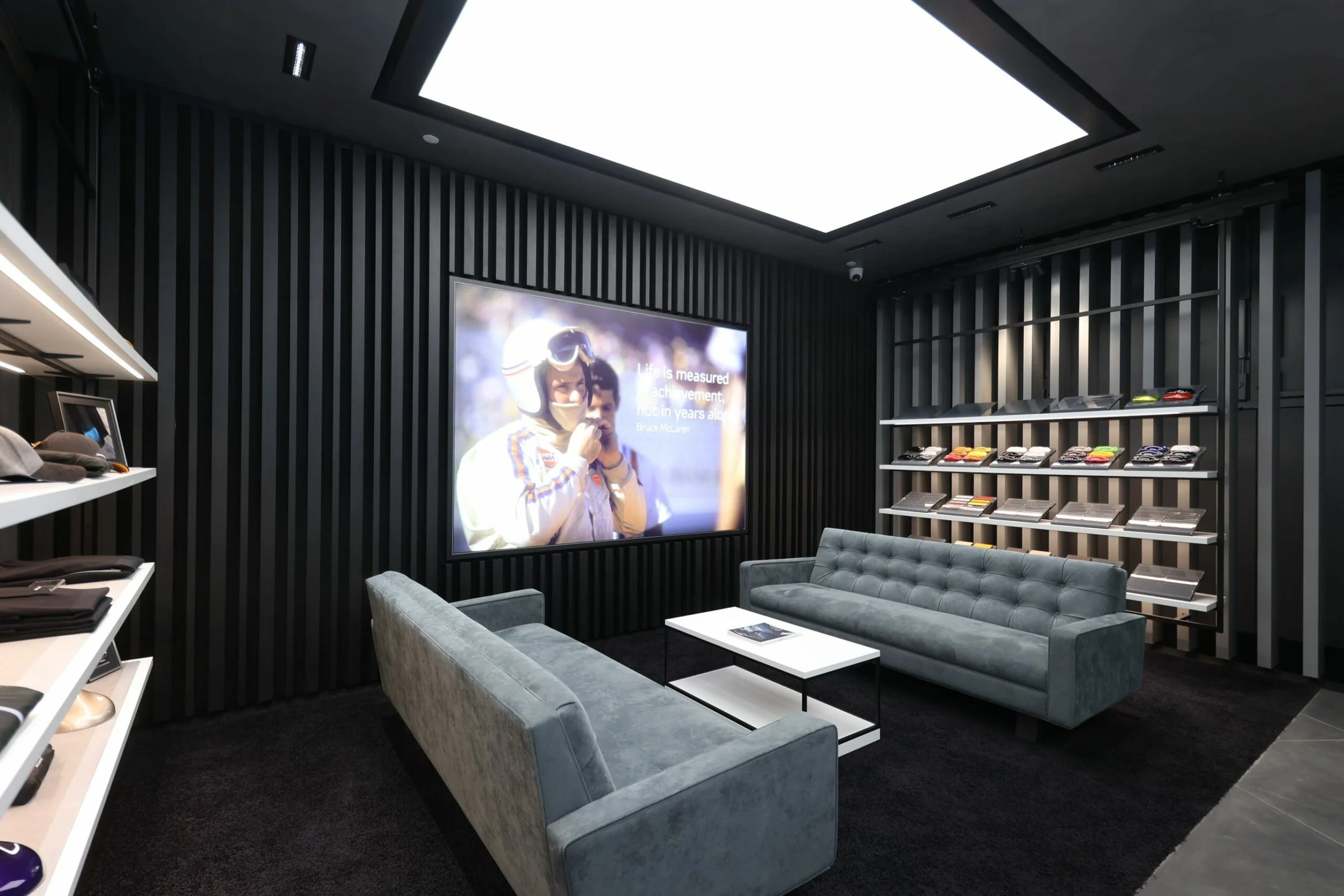 Interior of the McLaren showro with black striped wall paneling, a large illuminated ceiling panel, a display screen showing a person in racing gear, and a seating area with gray upholstered couches. There are shelves with hats, shoes, and magazines.
