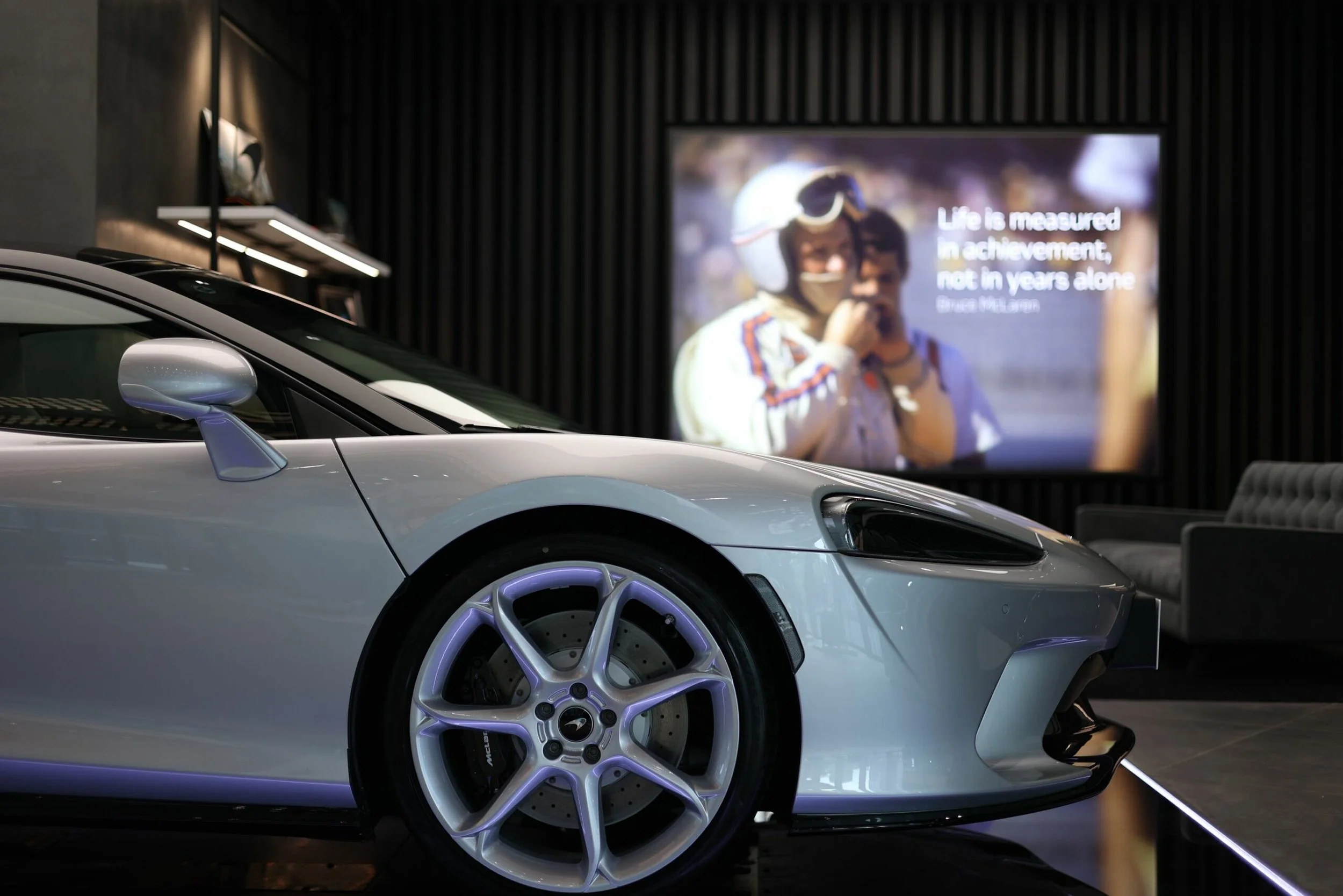 Close-up of a silver sports car with purple-accented wheels parked indoors. In the background, a large screen displays a woman and man with the quote, 'Life is measured in achievement, not in years alone' by Bruce Millan.