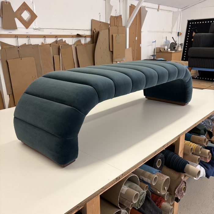 A modern, curved teal velvet Fleure bench with fluted cushions on a white worktable in a furniture workshop.