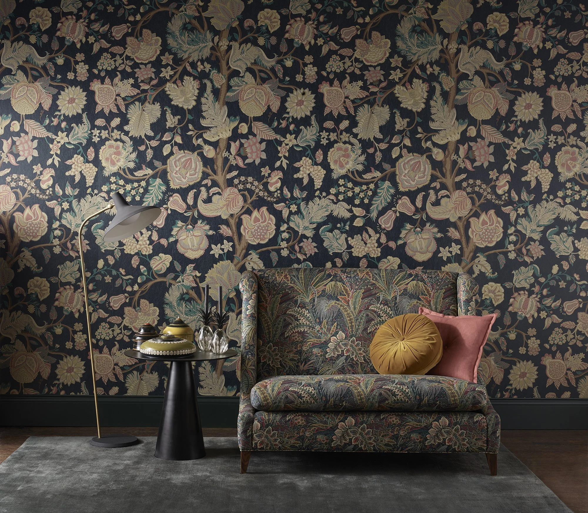 A dark floral patterned wall with a gold-colored velvet cushion and a floral upholstered bench in front. A black round side table holds decorative vases, and a white floor lamp is beside the wall.