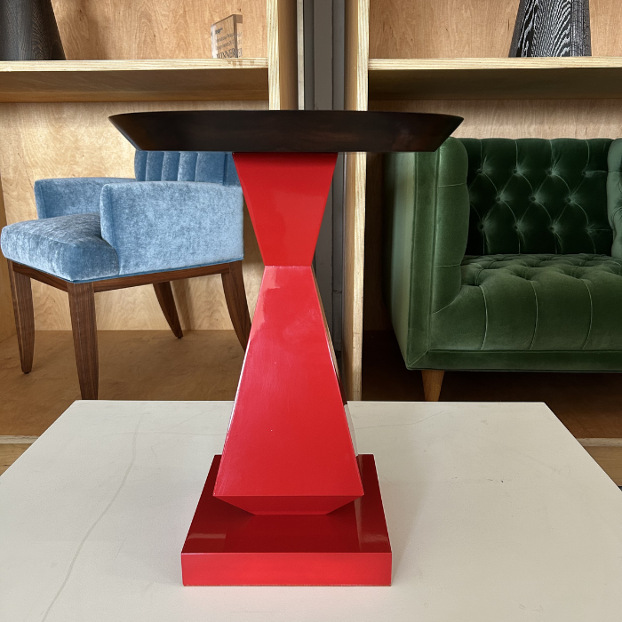 A modern side table with a black top and a red, geometric, sculptural base, placed on a white surface with two upholstered chairs in the background.