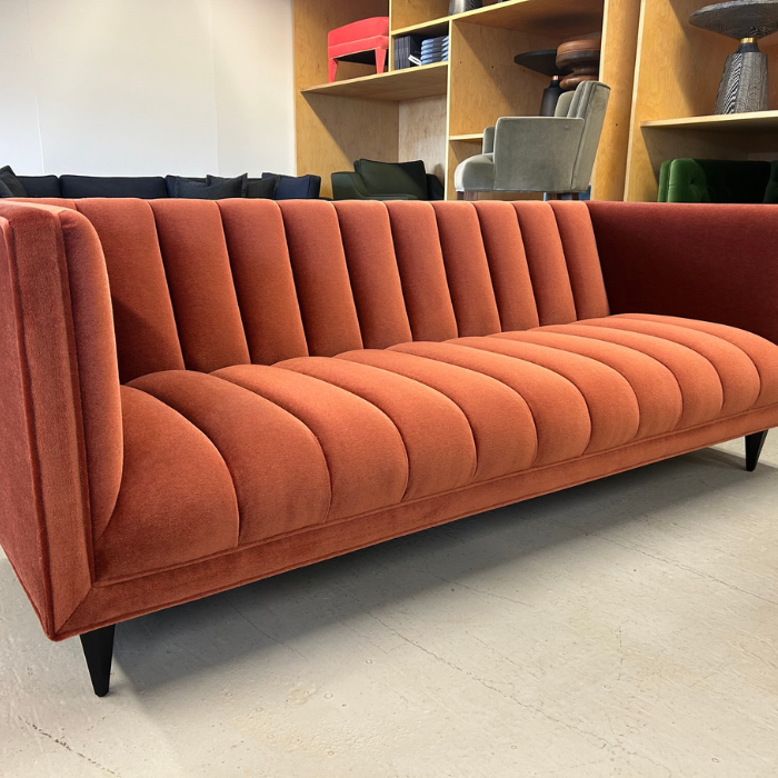 A coral-colored velvet sofa with vertical channel tufting, situated on a light tiled floor, with a background of modern chairs and wooden shelving units.