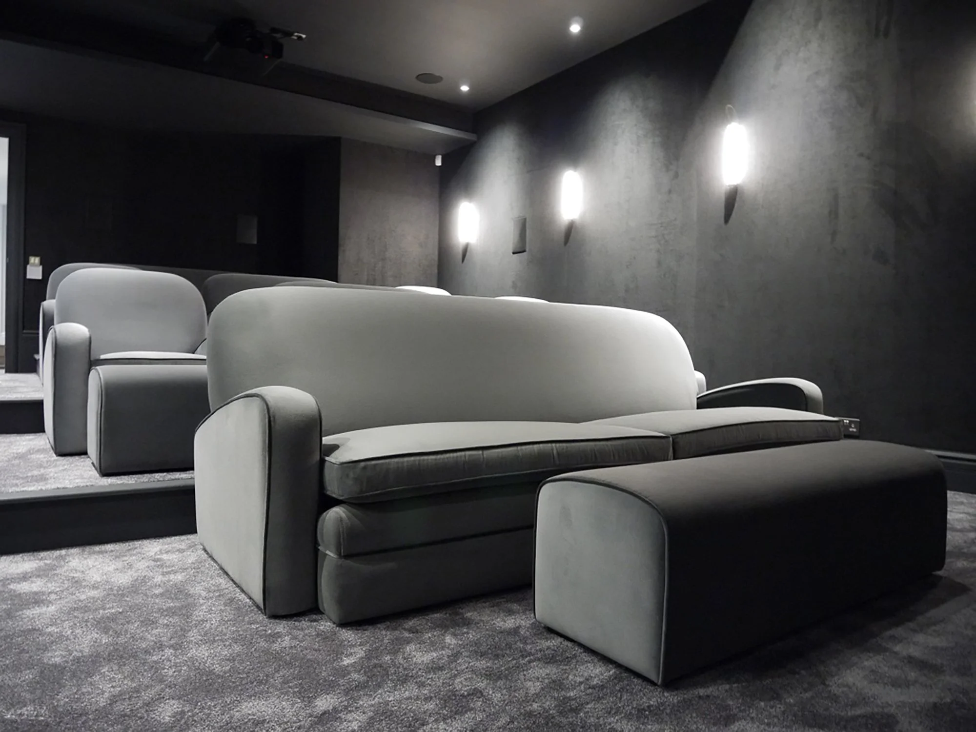 A row of light gray and dark gray plush armchairs arranged in a modern, dimly lit home cinema with dark walls and ceiling, featuring three wall-mounted light fixtures and a projector on the ceiling.