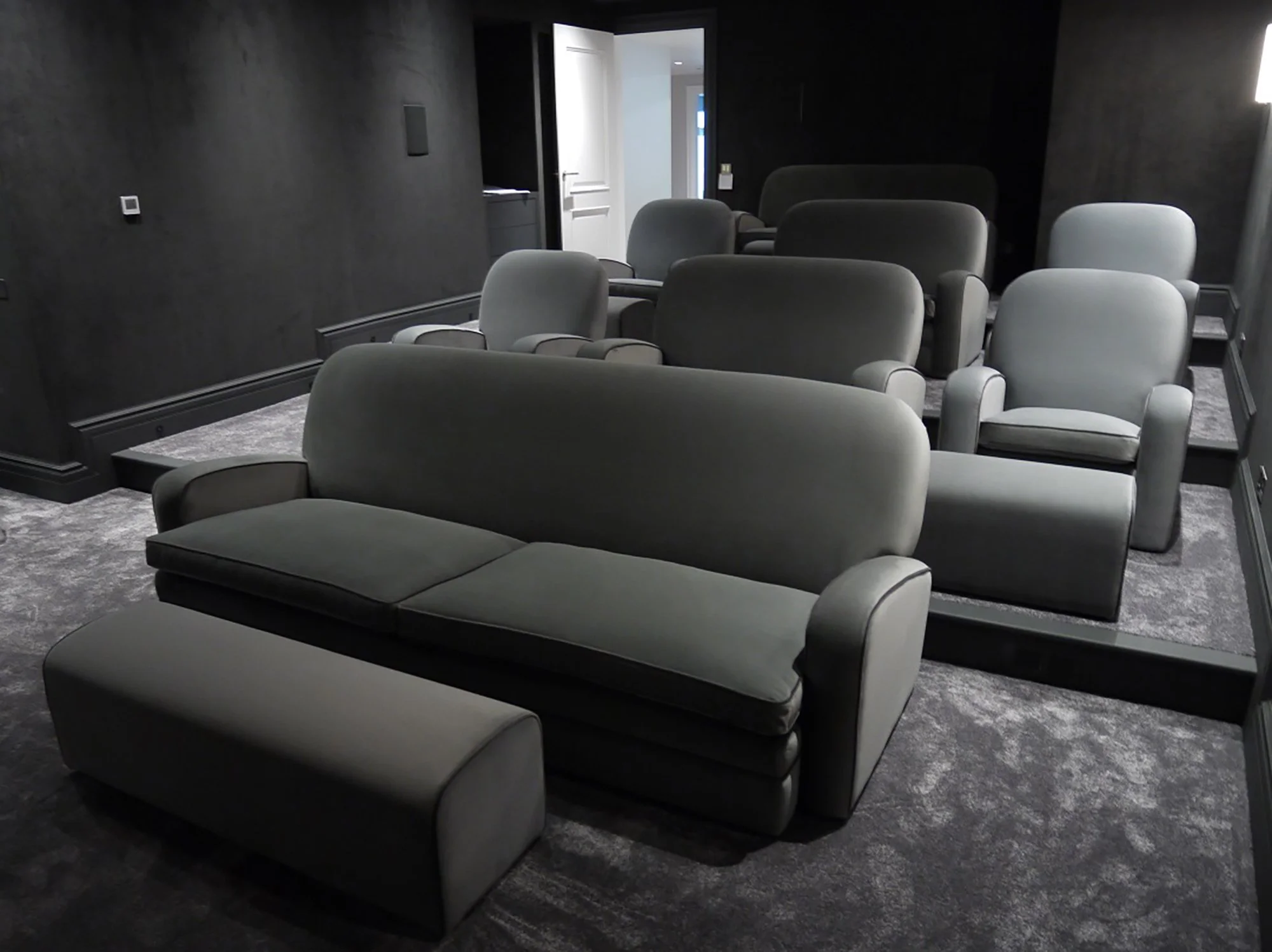 A dimly lit home cinema with grey upholstered chairs arranged in rows, grey walls, a carpeted floor, and an open door leading to a brighter space.