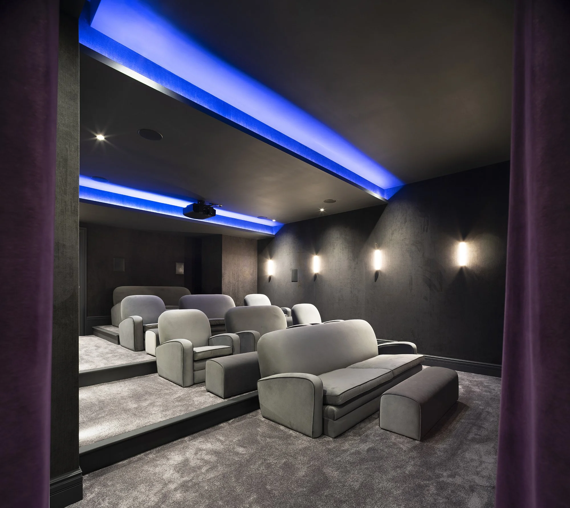 A modern, dimly lit home cinema with gray seating arranged in rows. The ceiling features blue accent lighting along the edges, and there is a ceiling-mounted projector. The walls are dark-colored, and purple curtains are visible on the sides.