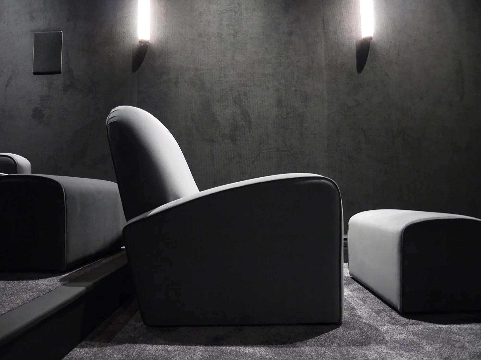 Empty gray theater with gray chairs and gray walls, illuminated by wall-mounted lights.