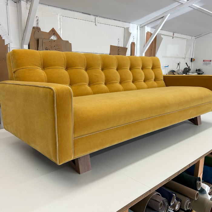 A vintage mustard yellow upholstered sofa with tufted backrest, placed in a workshop or storage area, with cardboard and workshop tools in the background.