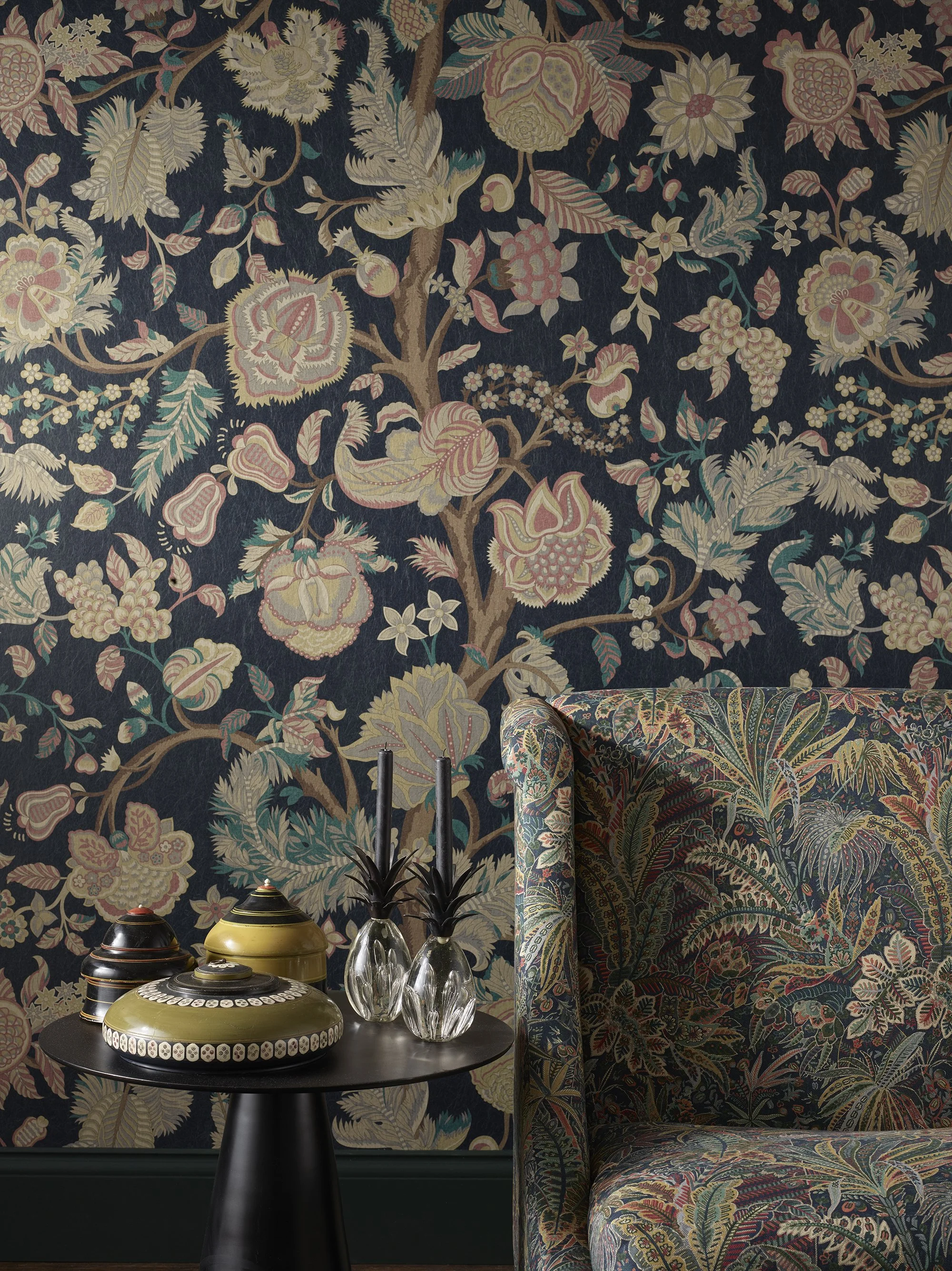A living room corner with a dark floral wallpaper featuring large, colorful flowers and leaves. A patterned armchair with a similar floral design is placed next to a black round side table. On the table, there are decorative vases and a ceramic object with black and yellow stripes. The background is dark, contrasting with the intricate and vibrant floral patterns.