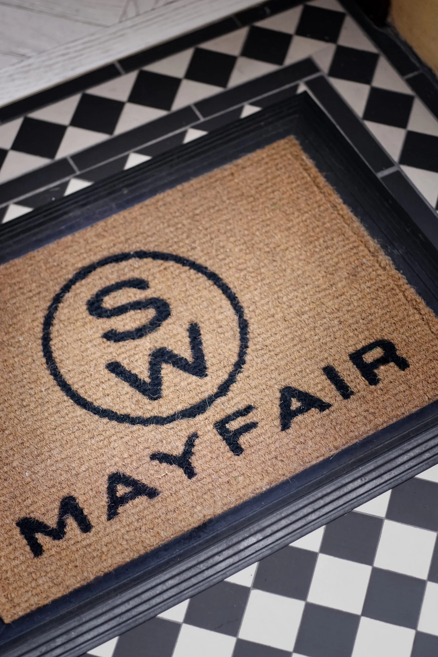 A doormat with the words 'WELCOME YAY' and a smiley face design on it, placed on a black rubber mat tread, with a black and white checkered floor visible in the background.