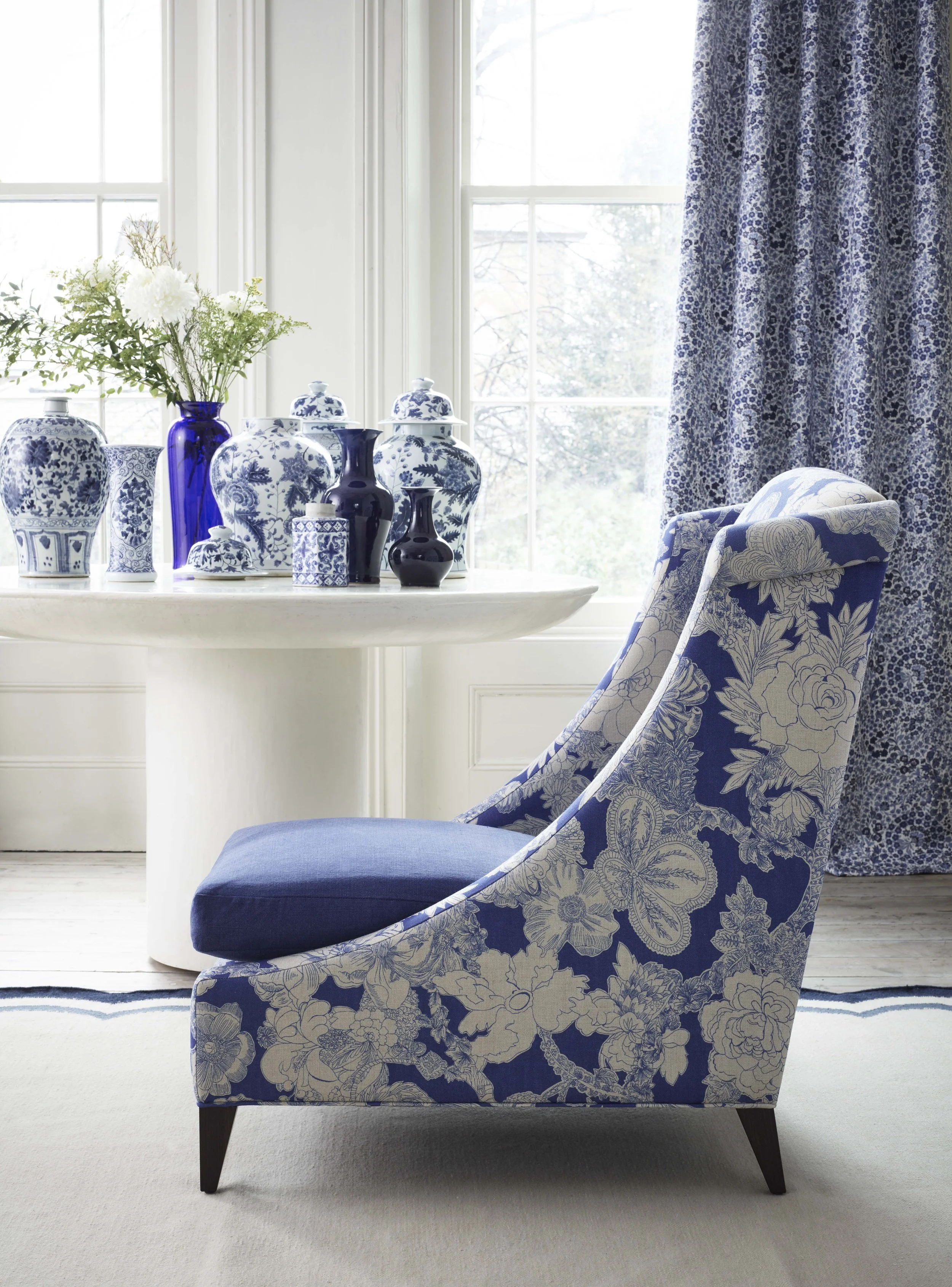 A living room corner with Liberty London blue floral fabric upholstered Vegas Lounger armchair, a white side table, and a window with patterned curtains. The side table holds blue and white decorative vases and a floral arrangement.