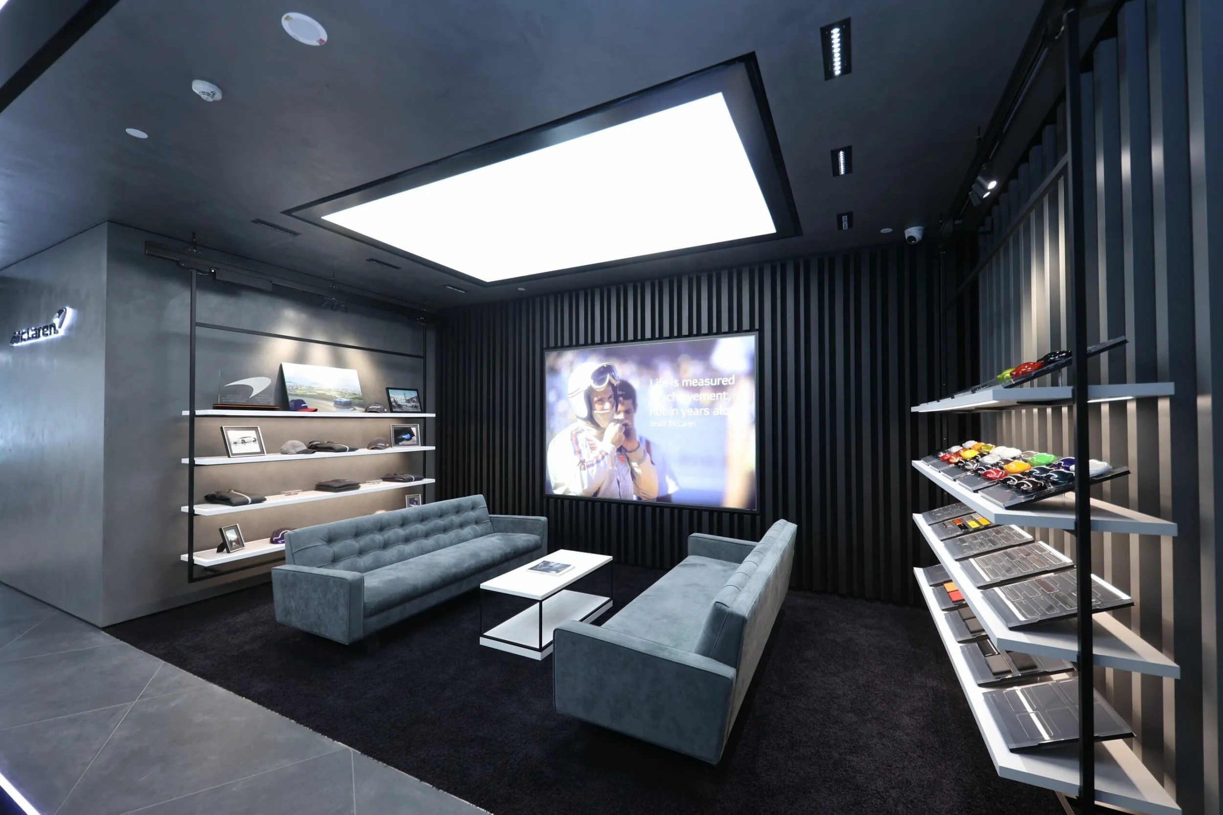 Interior of the McLaren showroom with a large bright ceiling light, grey walls, shelving with sunglasses and accessories, and a display screen showing a person at an event.